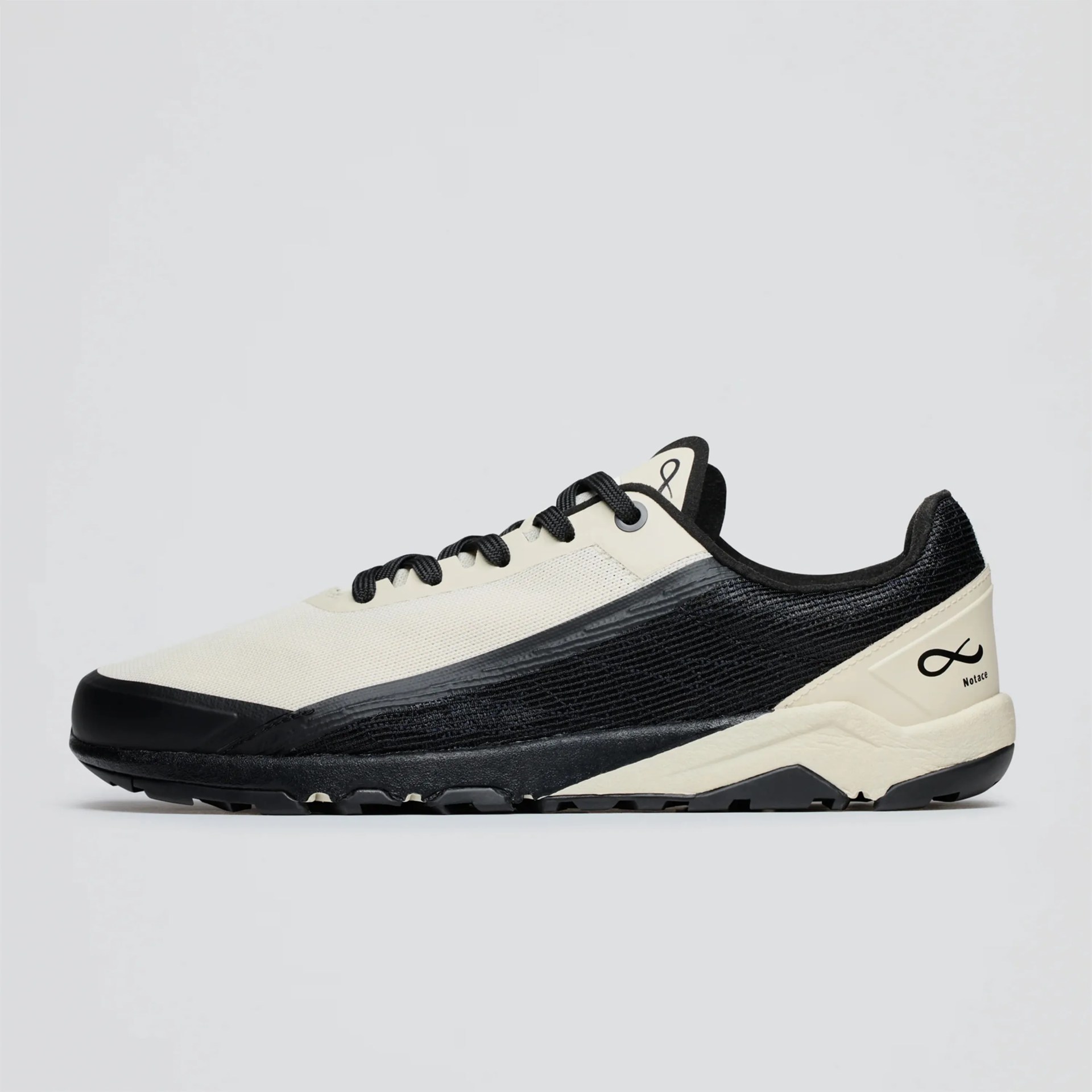 Low-profile sneaker with a sleek design featuring a cream-colored upper and black accents. The shoe has black laces, a black textured side panel, and a black sole with a rugged tread. The heel area includes a cream section with a black infinity symbol and the word "Notace" printed below it. The tongue also displays the infinity symbol.