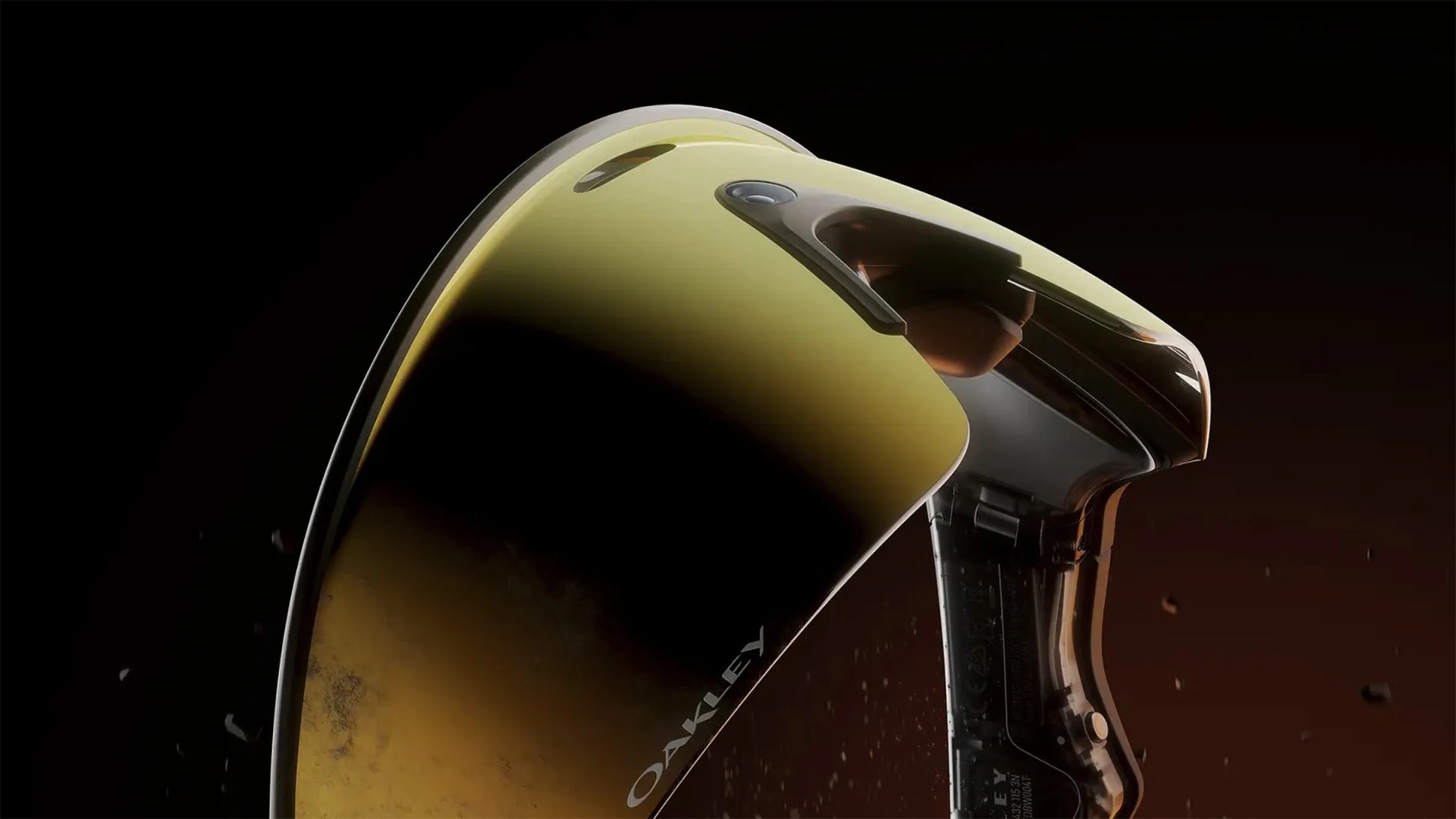 Close-up of a pair of Oakley sunglasses with a reflective yellow-gold lens and a transparent black frame, set against a dark background. The Oakley logo is visible on the lens.
