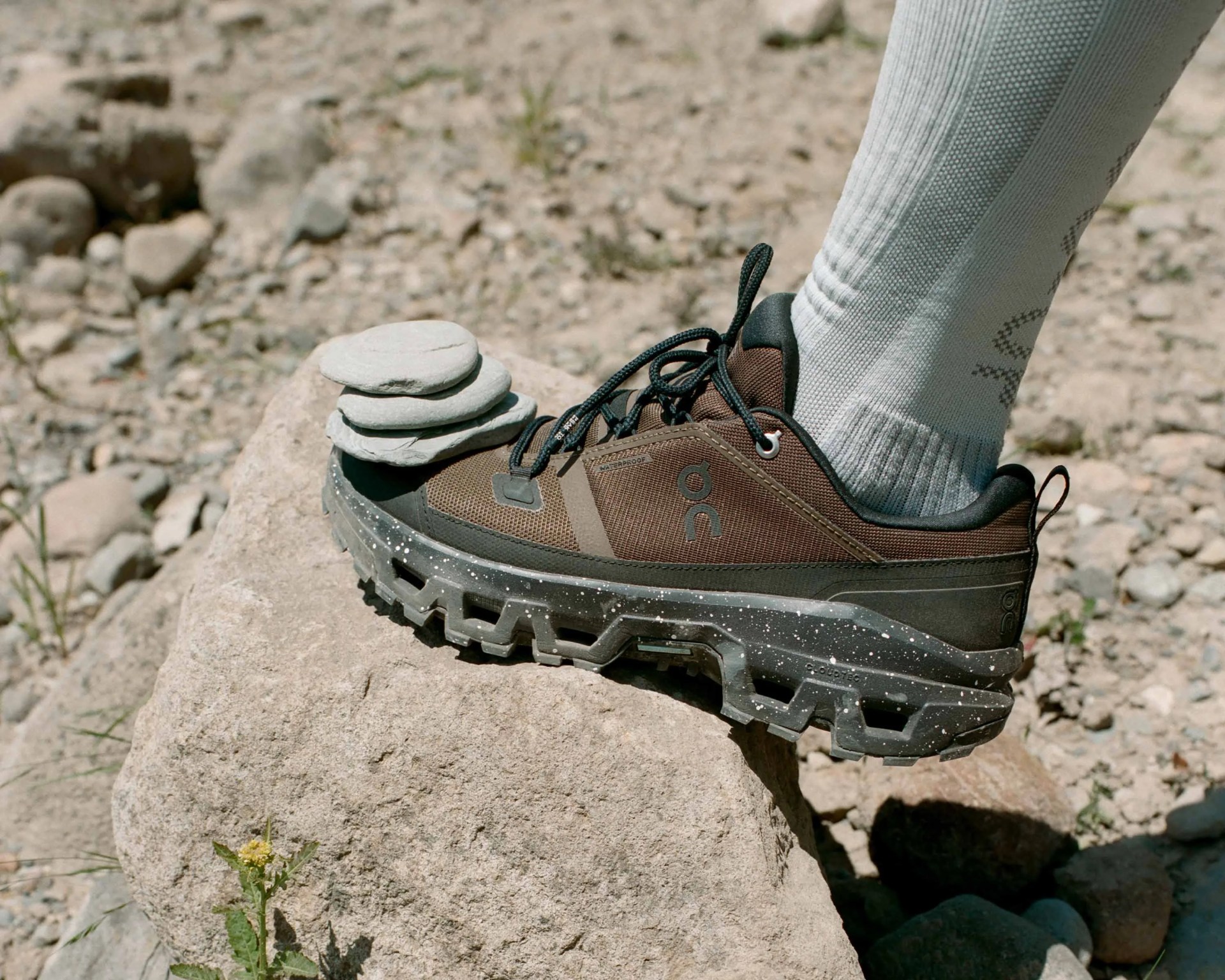 Brown and black hiking shoe with black laces and a rugged, speckled sole, worn with light gray socks, resting on a large rock outdoors with three flat stones stacked on the shoe's toe.