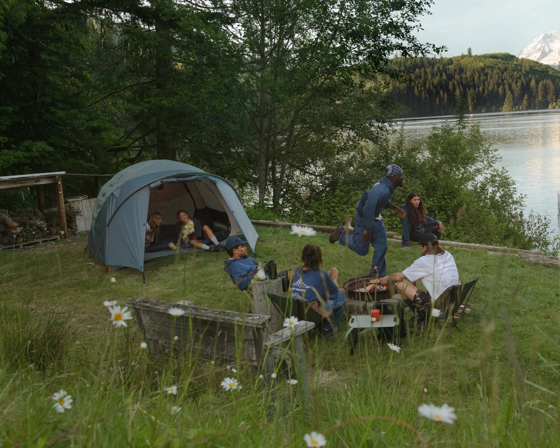 A group of six people camping by a lakeside, surrounded by trees and mountains. Two people are sitting inside a blue tent, while the others are gathered around a fire pit on chairs and a wooden bench. Wildflowers and tall grass are in the foreground, and the lake and forested hills are visible in the background.