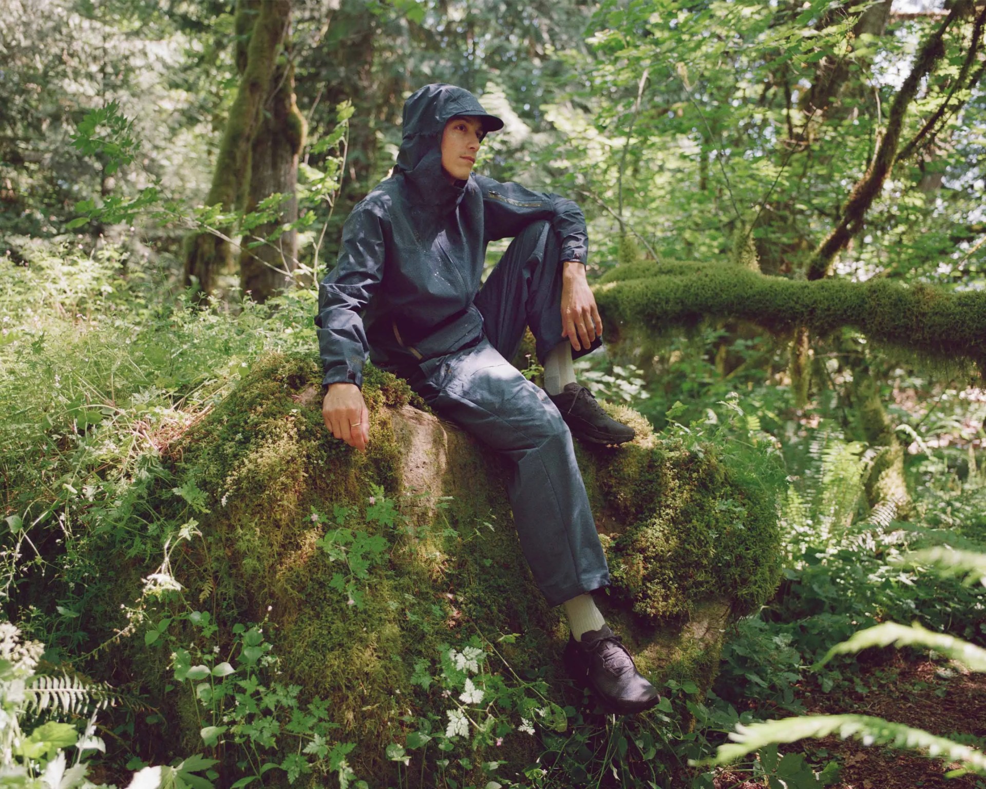 A person wearing a dark blue rain jacket with a hood, matching pants, white socks, and black shoes is sitting on a large moss-covered rock in a lush green forest. The individual is looking thoughtfully into the distance, surrounded by dense foliage and trees.