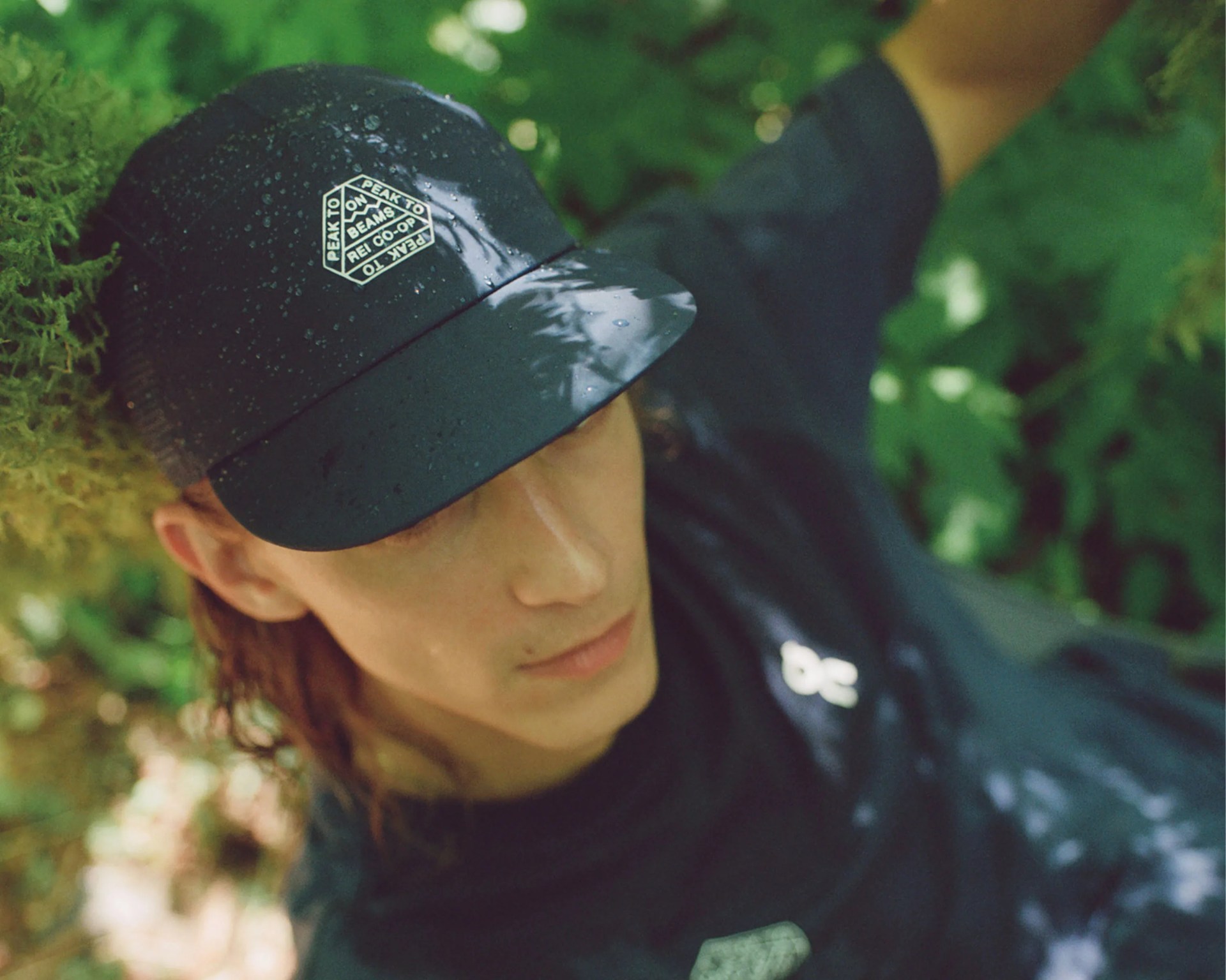 A person wearing a black cap with water droplets on it, featuring a white hexagonal logo that reads "PEAK TO PEAK TO PEAK TO ON BEAMS REL CO-OP." The person is also wearing a dark shirt with a small white logo on the chest, surrounded by green foliage.