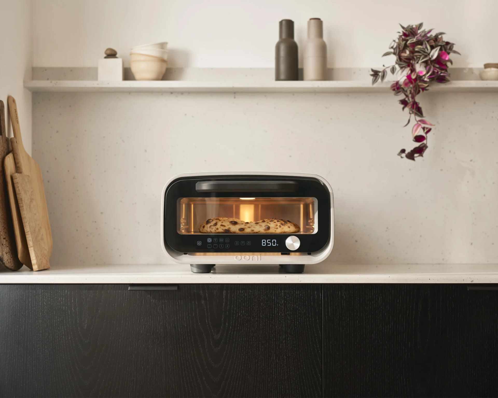 A modern countertop pizza oven with a digital display showing "850" and a silver control knob, baking a pizza inside. The oven is placed on a white countertop with dark wood cabinets below. In the background, there is a white wall with a floating shelf holding two ceramic bottles, a small stack of bowls, and a hanging plant with purple and green leaves. On the left side of the countertop, several wooden cutting boards are leaning against the wall.