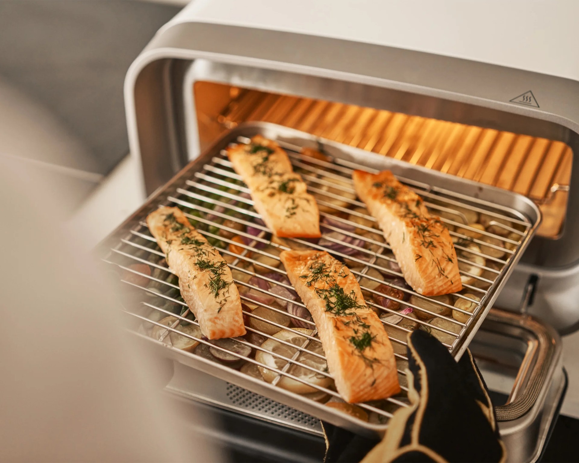 Four seasoned salmon fillets are being taken out of a small oven on a metal rack, with sliced vegetables underneath. A person wearing a black oven mitt with beige trim is holding the rack. The oven interior is illuminated with a warm light.