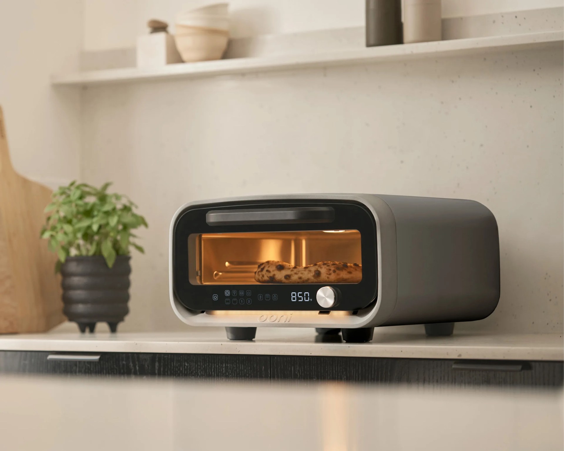 A modern countertop pizza oven with a digital display showing 850°F, baking a pizza inside. The oven is gray with a black front panel and a silver control knob. It is placed on a light-colored kitchen countertop with a potted green plant and wooden cutting board in the background.