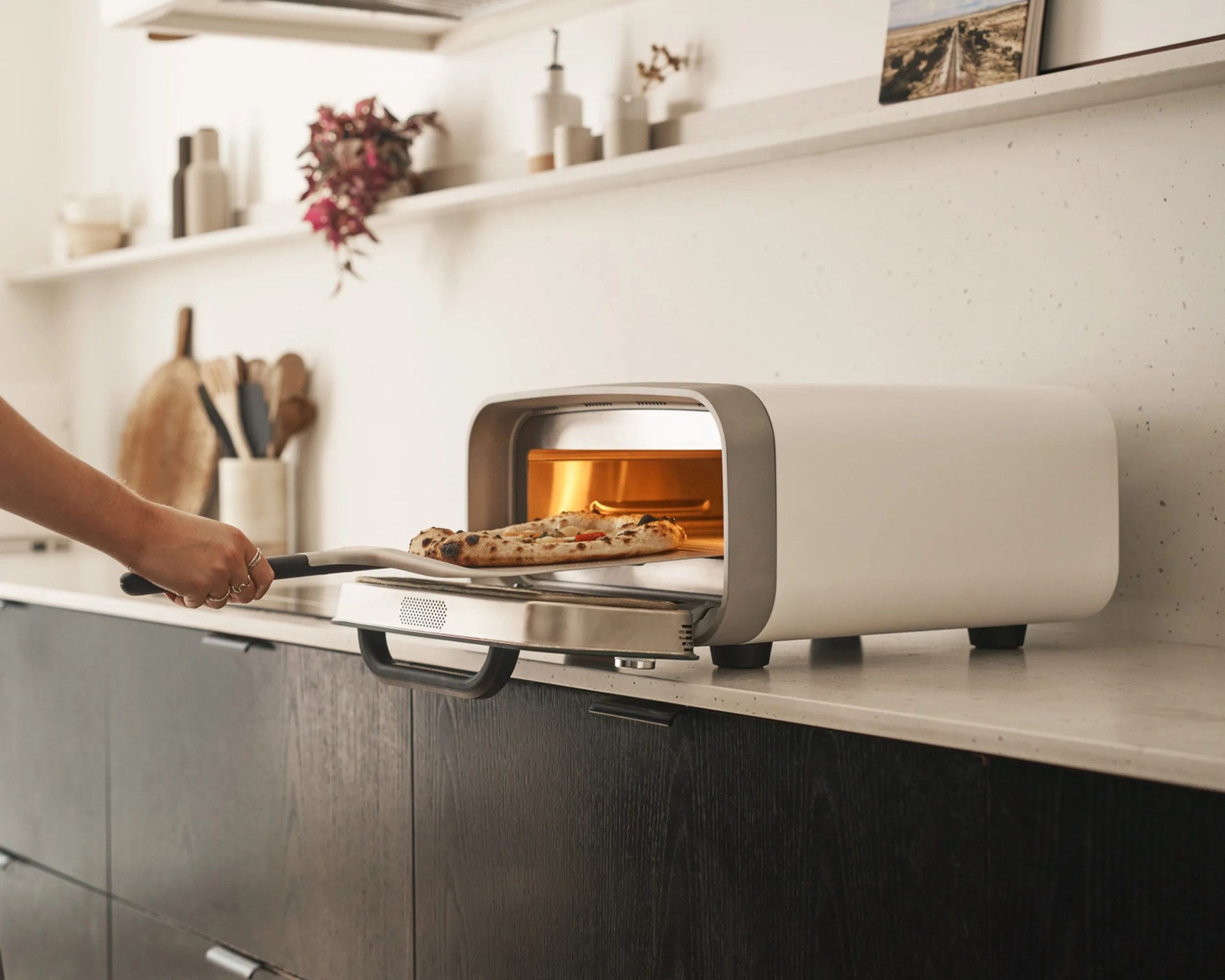 A modern countertop pizza oven with a white and gray exterior is shown on a kitchen counter. A hand is using a pizza peel to remove a freshly baked pizza with a golden, slightly charred crust from the oven. The kitchen has dark wood cabinets, a light-colored countertop, and a white speckled backsplash. Various kitchen utensils and decor items are visible in the background.