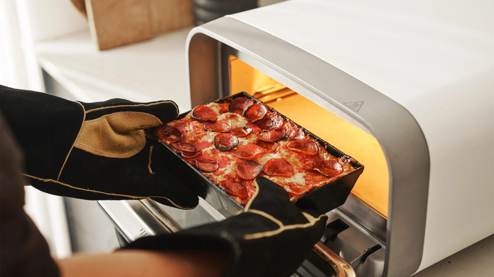 A person wearing black and tan oven mitts is removing a rectangular pepperoni pizza from a small white countertop oven. The pizza has melted cheese and browned pepperoni slices. The oven interior is illuminated with a warm light.