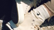 A close-up of a person wearing off-white high-top canvas sneakers with metal eyelets and white ribbed socks featuring a black and white yin-yang symbol. The person is also wearing light brown pants with frayed hems. The background appears to be a textured outdoor surface.