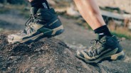 A close-up of a person wearing black and beige Salomon trail running shoes with black socks, stepping on a rocky, uneven outdoor trail. The shoes appear sturdy and designed for rugged terrain.