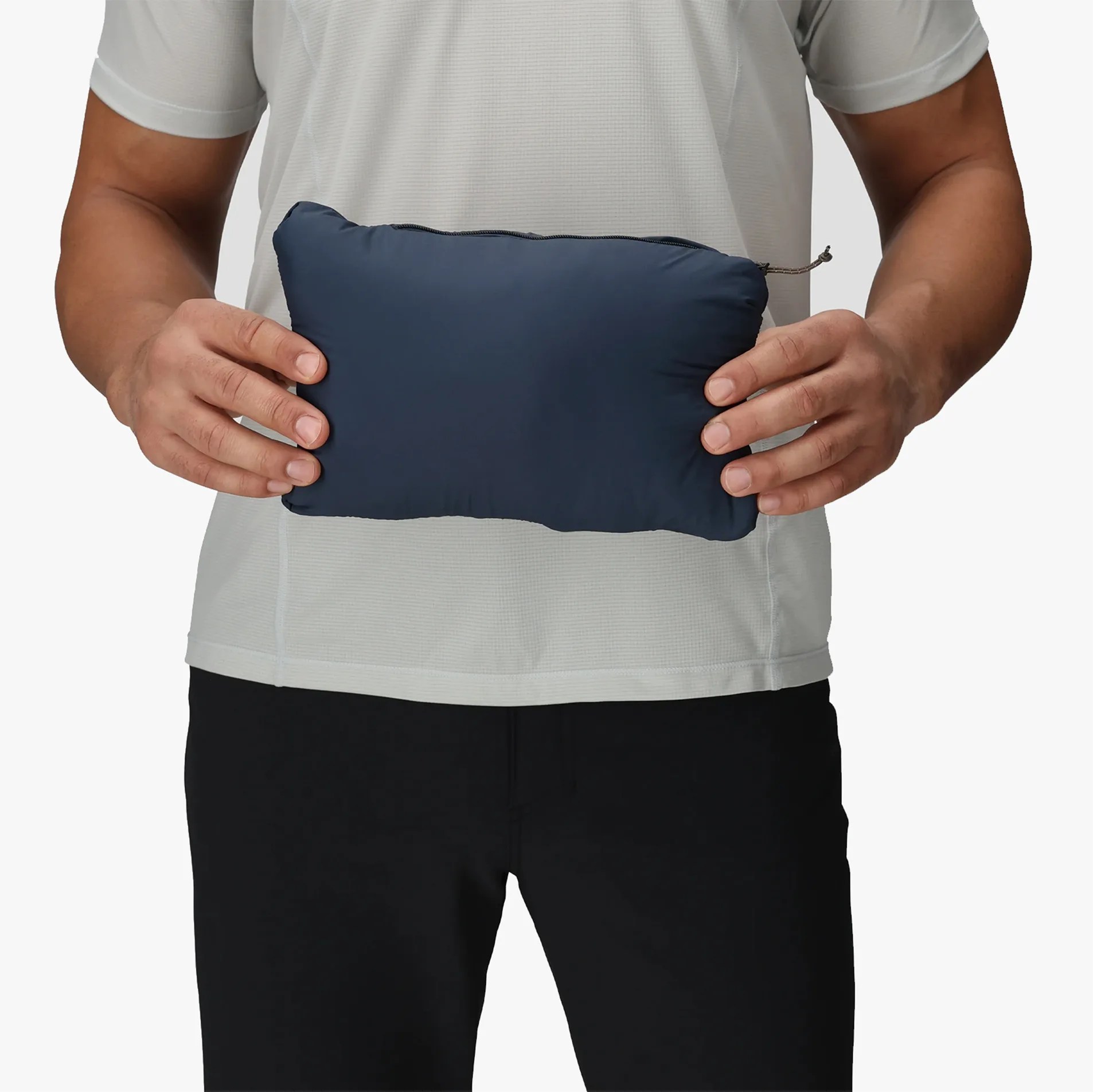 A person wearing a light gray short-sleeve shirt and black pants is holding a small, rectangular, navy blue zippered pouch with both hands at waist level.