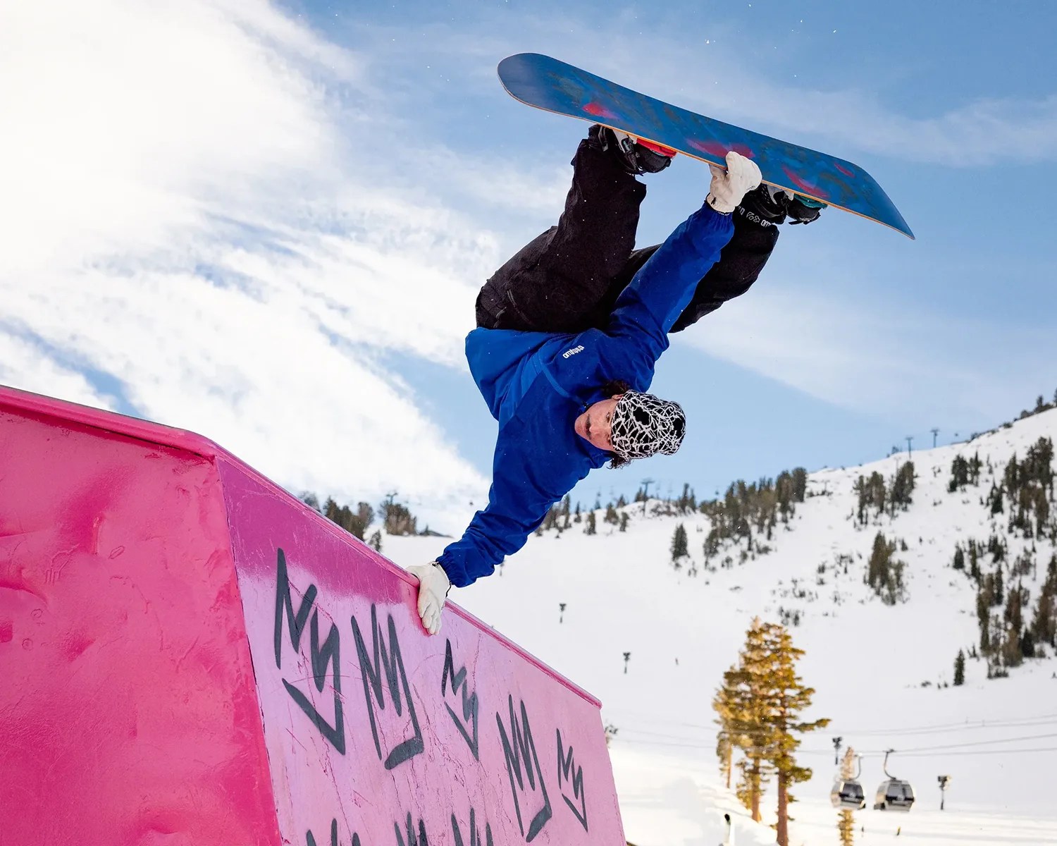 A snowboarder wearing a blue jacket, black pants, white gloves, and a black-and-white patterned beanie is performing a handplant trick on a bright pink rail with black crown graffiti. Snow-covered mountains and trees are visible in the background under a partly cloudy blue sky.