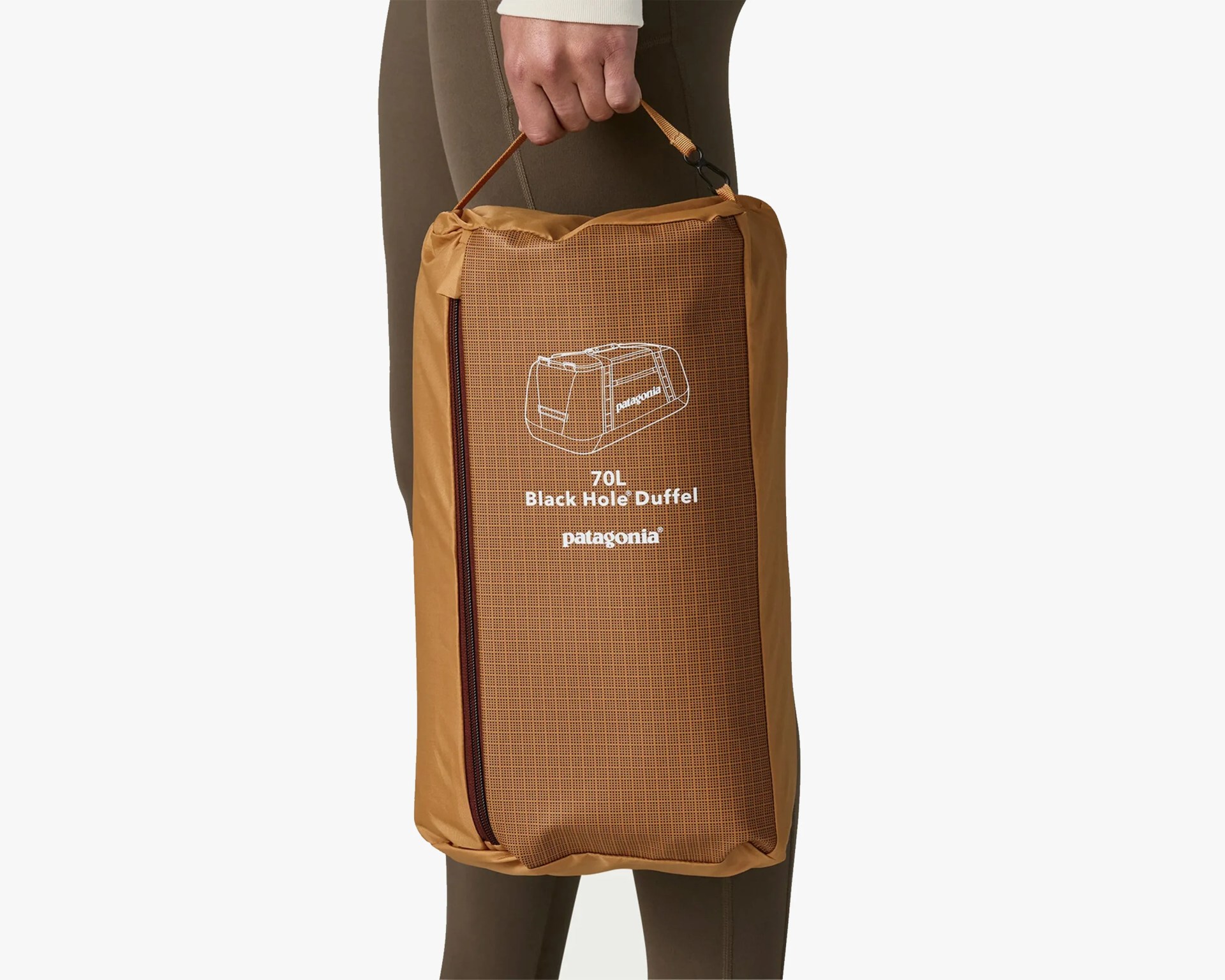 A person is holding a packed Patagonia 70L Black Hole Duffel bag in a brown storage sack with a zipper. The sack features a white outline illustration of the duffel bag and the text "70L Black Hole Duffel patagonia" on the front. The person is wearing olive green leggings and a light-colored long sleeve top.