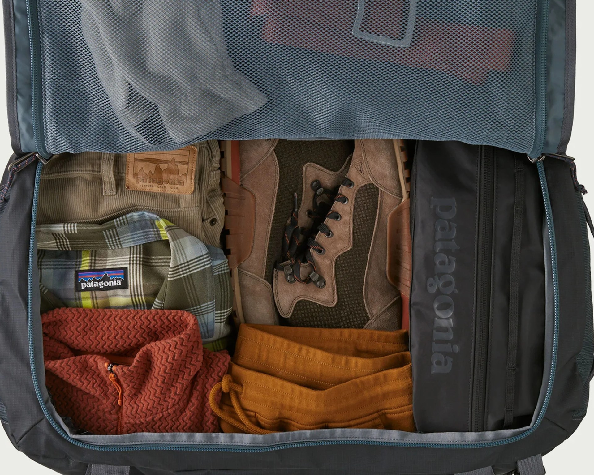 A packed suitcase with neatly folded clothing and hiking boots. Visible items include a green and brown plaid Patagonia shirt, tan corduroy pants with a Patagonia leather patch, rust-colored textured fleece, mustard-colored pants, brown hiking boots, and a black Patagonia zippered pouch. The suitcase has a mesh zippered compartment on the inside of the lid.