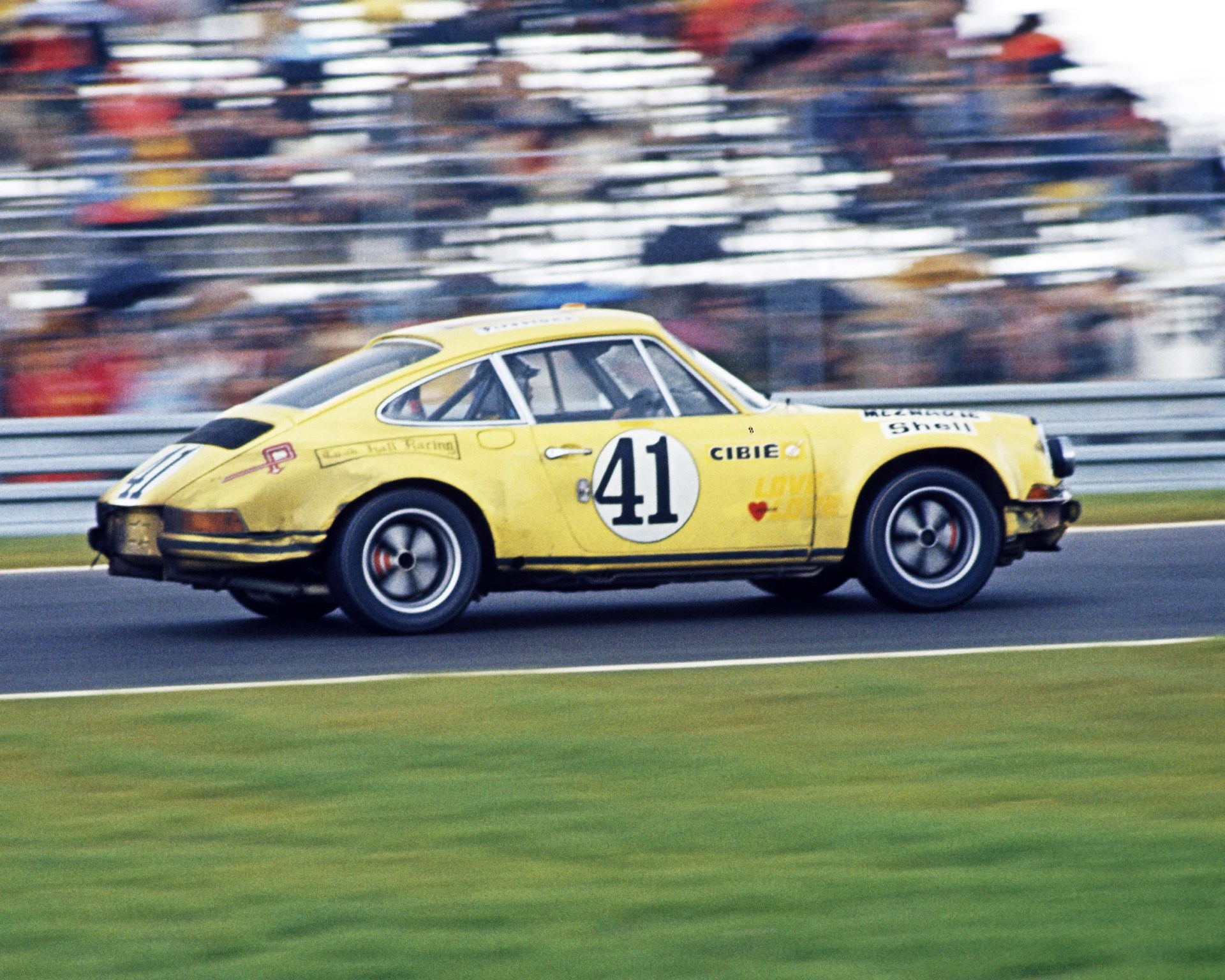 Yellow vintage race car number 41, with sponsor decals including "Cibie" and "Shell," driving on a racetrack with blurred spectators in the background.