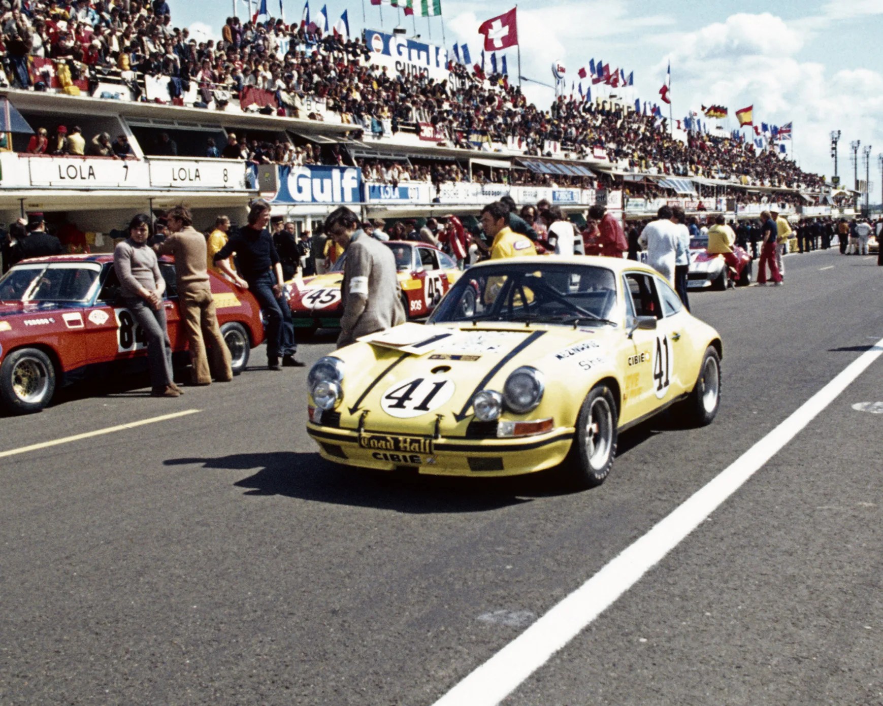 A yellow vintage race car with the number 41 on the hood and doors is parked on a race track pit lane. The car has black racing stripes and additional headlights mounted on the front. Behind it, several other race cars and people are visible, with a large crowd in the grandstands above. Flags from various countries are flying above the grandstands. The atmosphere suggests a classic motorsport event.