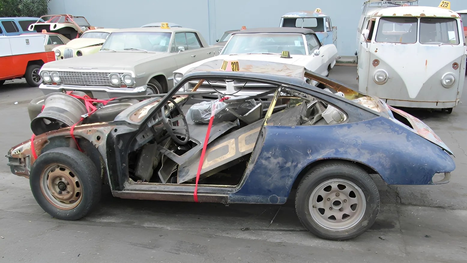 A stripped-down, partially disassembled blue vintage car with no doors or front fenders, filled with various car parts inside. The car is parked on a lot with other classic vehicles, including a beige car, a white convertible, and a white Volkswagen van in the background. The blue car's front wheel is rusty, and the rear wheel has a different rim. Red straps are securing some parts inside and on the car.