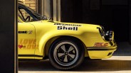 Yellow vintage race car front side with black and silver wheel, featuring sponsor decals including "Shell," "BIE," and "LOVE DESIGN" with a red heart. The car is partially inside a garage with sunlight illuminating the front.
