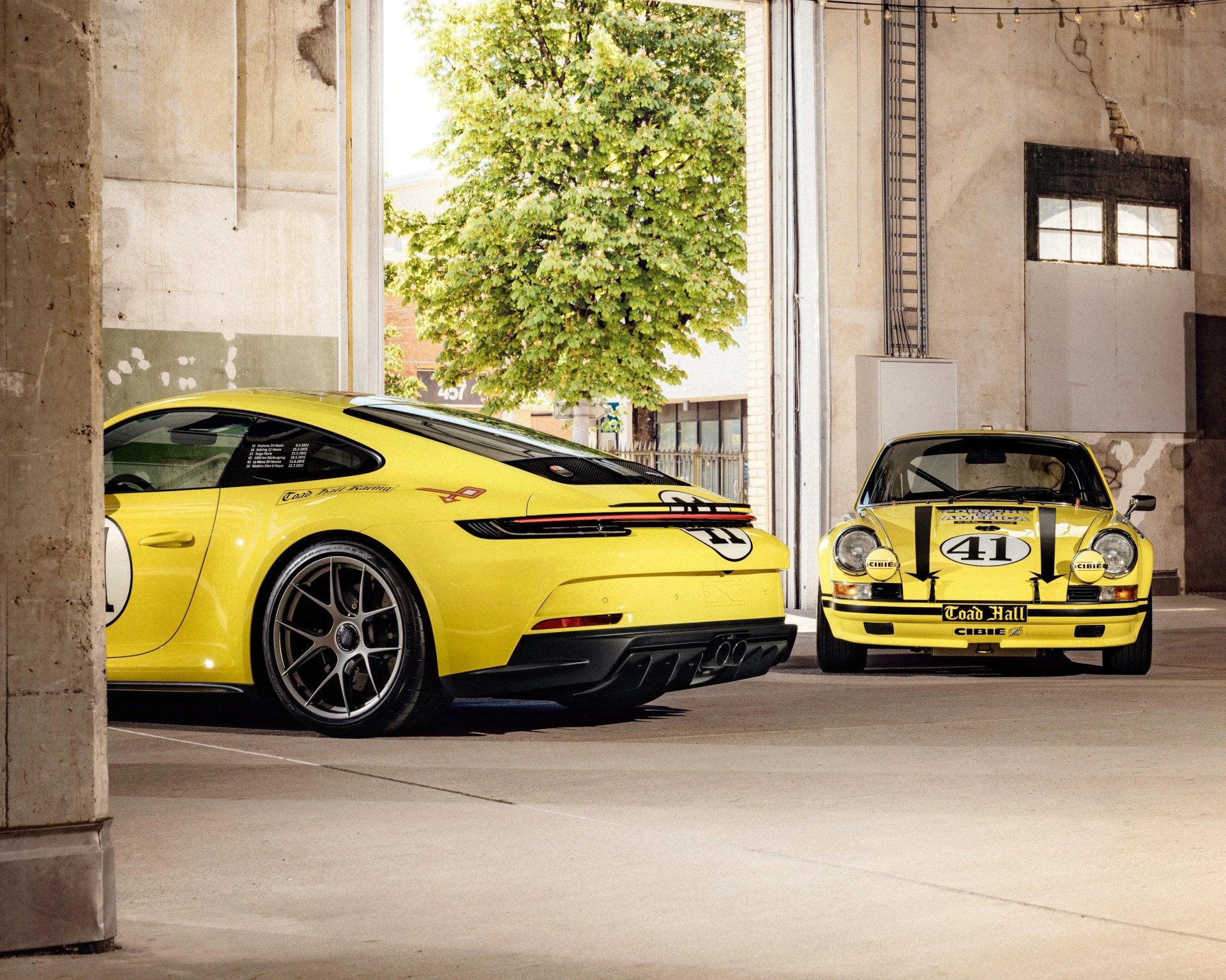 Two yellow Porsche sports cars are parked inside a concrete garage with an open door. The car on the left is a modern Porsche 911 with a sleek design, black wheels, and racing decals including the number 41 on the door. The car on the right is a classic Porsche 911 with round headlights, black racing stripes, and the number 41 on the hood. Both cars have "Toad Hall" and "Cibie" decals. A green leafy tree is visible outside through the open garage door.