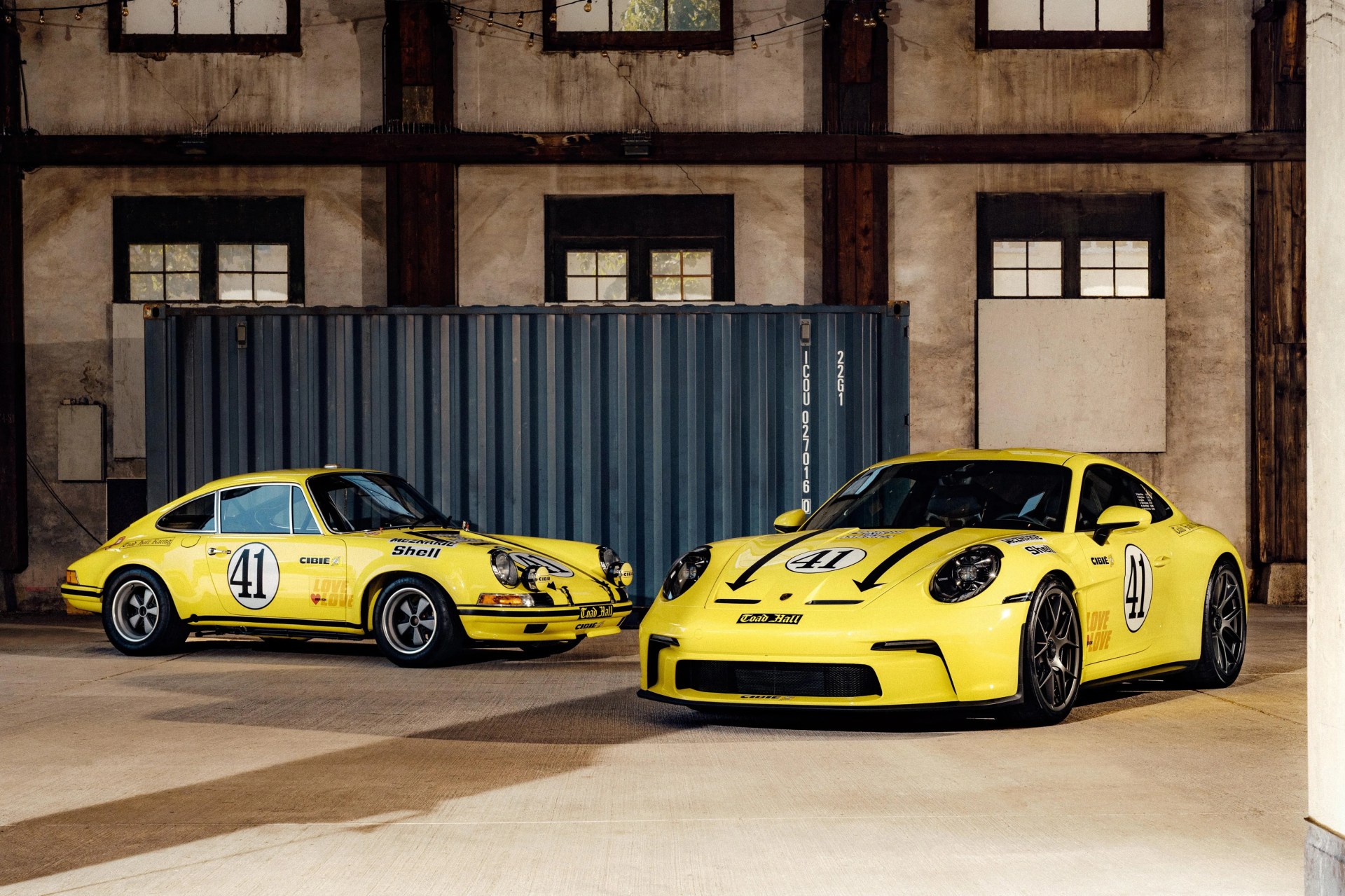 Two yellow Porsche sports cars with the number 41 on their doors and hoods are parked indoors in front of a large blue shipping container. The car on the left is a classic model with round headlights and additional rally lights on the front bumper. The car on the right is a modern model with sleek, aerodynamic lines and black racing stripes on the hood. Both cars have black wheels and various sponsor decals, including "Shell" and "Cibie." The setting is an industrial-style building with large windows and exposed wooden beams.