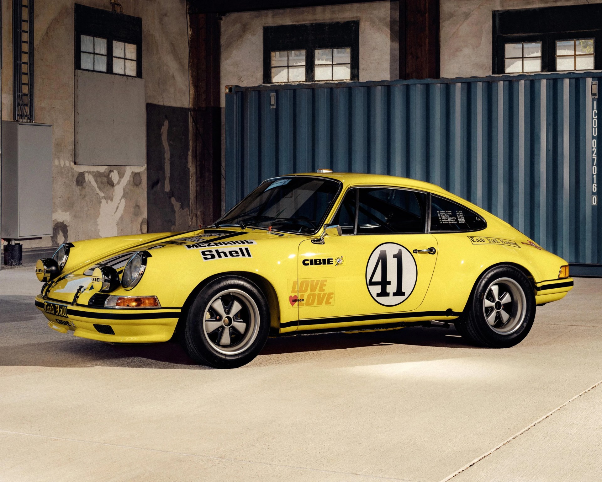 Yellow vintage Porsche 911 race car with black racing stripes, number 41 on the door, additional front-mounted rally lights, and sponsor decals including Shell and Cibie, parked indoors on a concrete floor with a blue shipping container and industrial walls in the background.