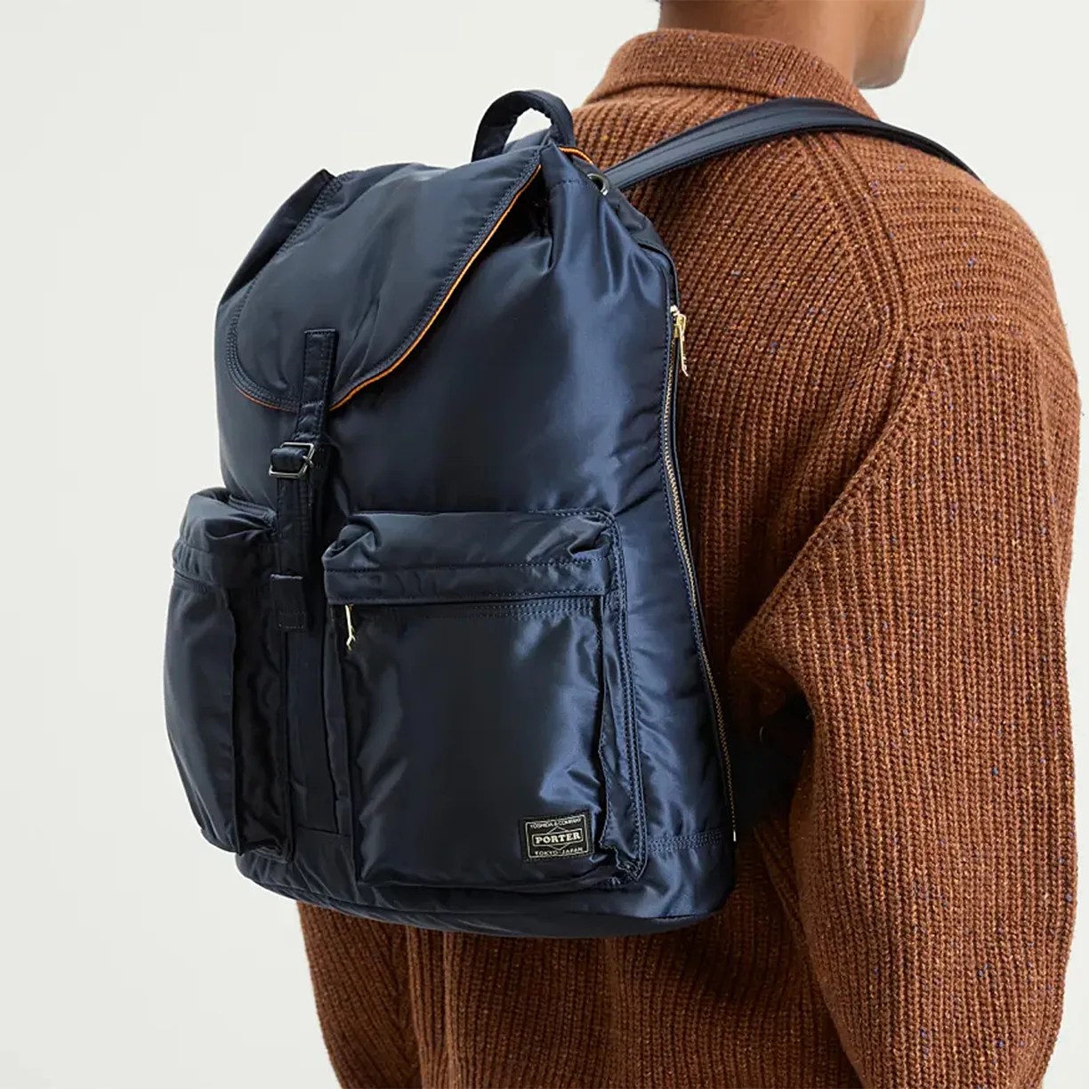 Navy blue backpack with multiple front pockets and a flap closure, worn over one shoulder by a person in a brown knit sweater. The backpack has a small rectangular label near the bottom right corner.