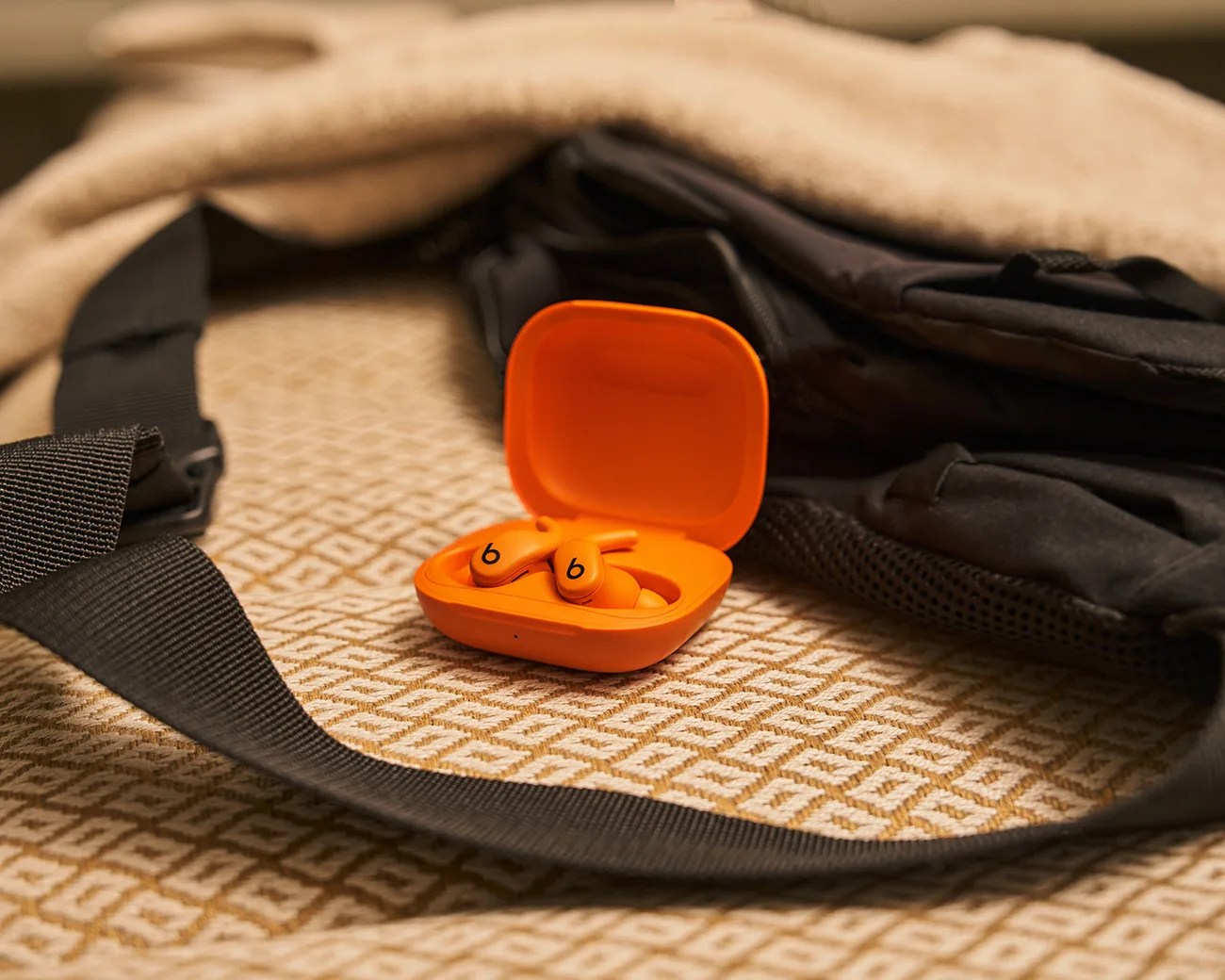 Bright orange wireless earbuds with a matching open charging case resting on a textured beige surface, next to a black fabric item with a strap.