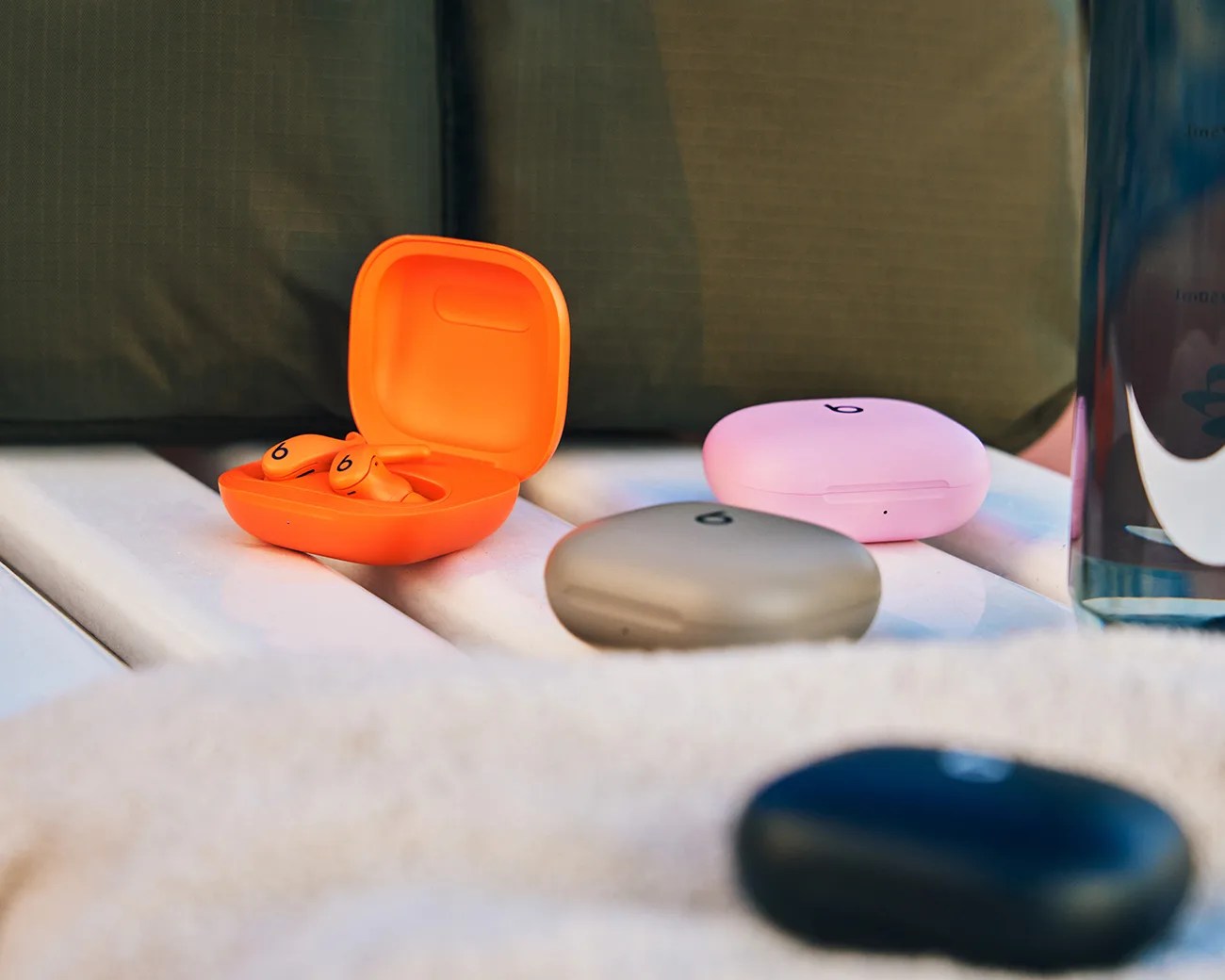Three pairs of wireless earbuds in their charging cases are placed on a white surface. One case is open and orange, revealing matching orange earbuds inside. The other two cases are closed, one pink and one beige. A dark blue object is partially visible in the foreground, and a green cushion is in the background.