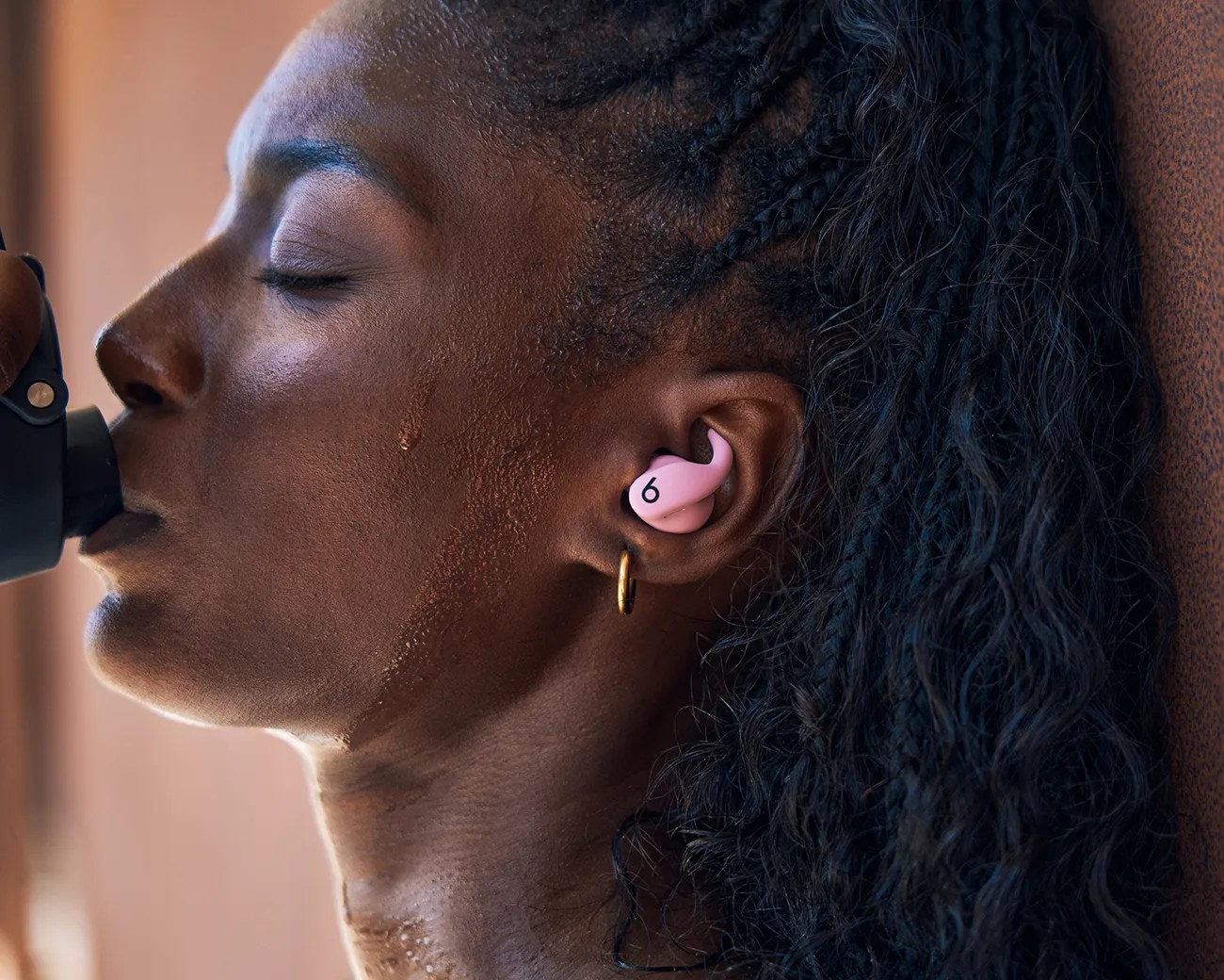 Close-up of a person with braided hair wearing pink wireless earbuds and a small gold hoop earring, drinking from a black water bottle with eyes closed and sweat visible on their face and neck.