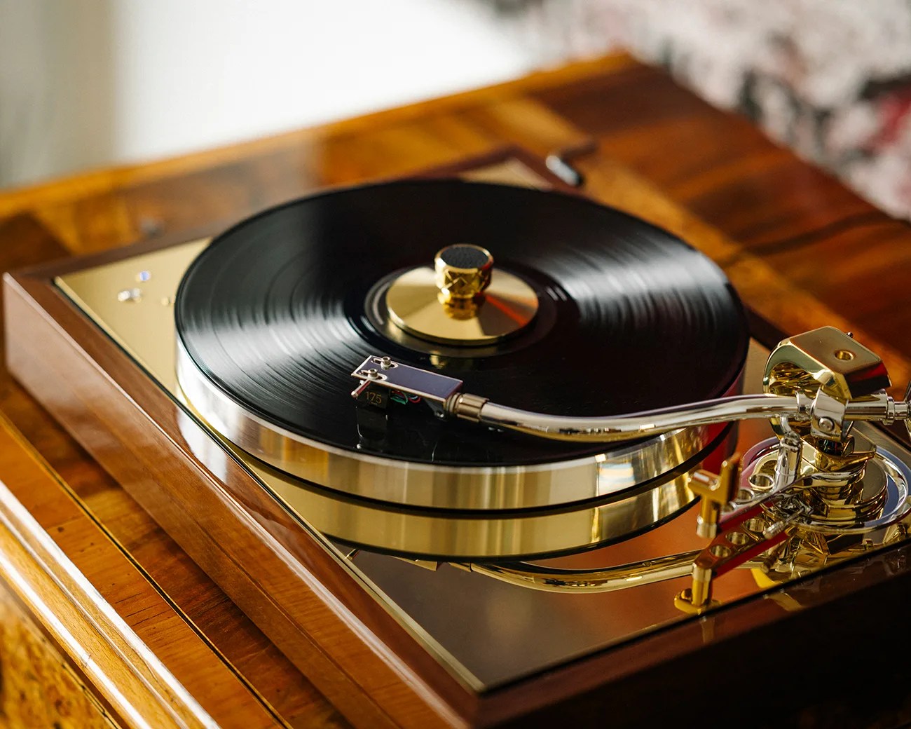 A close-up of a vintage turntable with a black vinyl record on it. The turntable features a polished wooden base and shiny gold and silver metal components, including the tonearm and record clamp. The surface beneath the record is reflective, adding to the luxurious appearance.