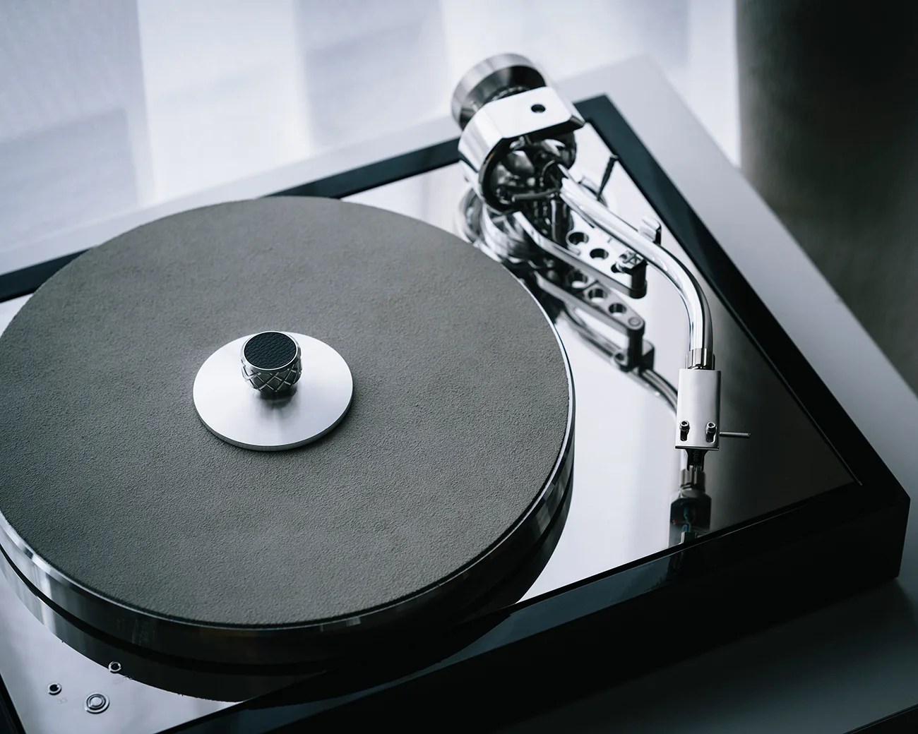 A close-up of a modern turntable with a sleek, reflective black base, a textured gray platter mat, and a shiny silver tonearm. The turntable has a minimalist design with a central spindle featuring a knurled metal knob.