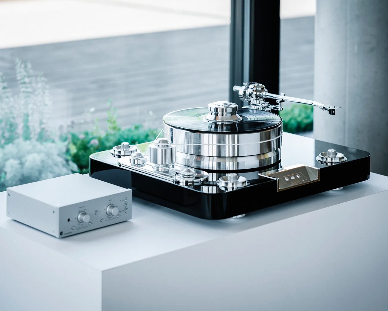 A modern, high-end turntable with a sleek black base and multiple shiny metallic components, including a thick platter and a tonearm. It is placed on a white surface next to a small silver control box with knobs and switches. The background shows a window with blurred greenery outside.