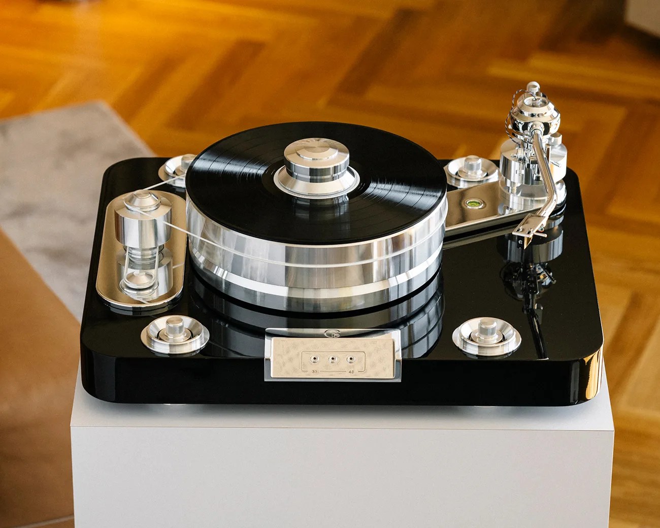 High-end turntable with a sleek black base and polished silver components, including a large platter holding a black vinyl record, a tonearm with a cartridge, and multiple control knobs and mechanisms, placed on a white pedestal against a wooden floor background.