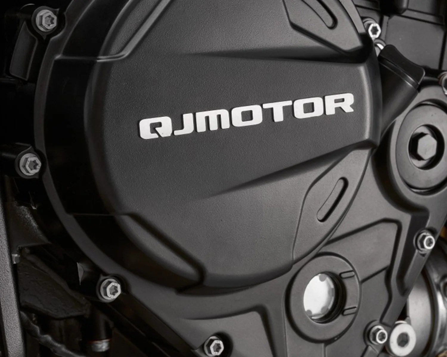 Close-up of a black motorcycle engine cover with the brand name "QJMOTOR" embossed in white letters. The cover is secured with multiple silver bolts.