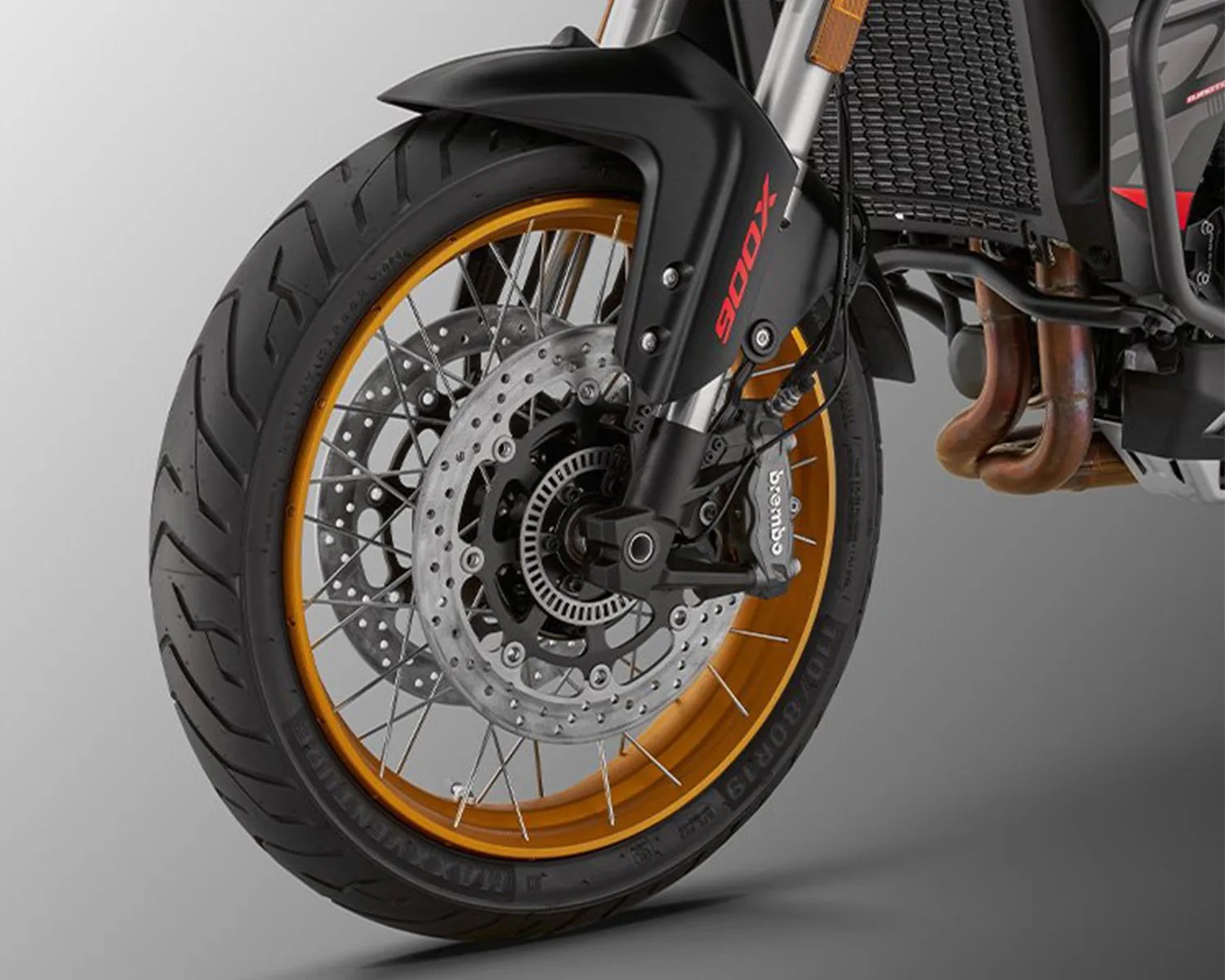 Front wheel of a motorcycle featuring a black tire with a gold rim, silver spokes, dual disc brakes, and a black brake caliper labeled "Brembo." The black front fender has "900X" written in red. Part of the motorcycle's exhaust pipes and radiator are visible in the background.