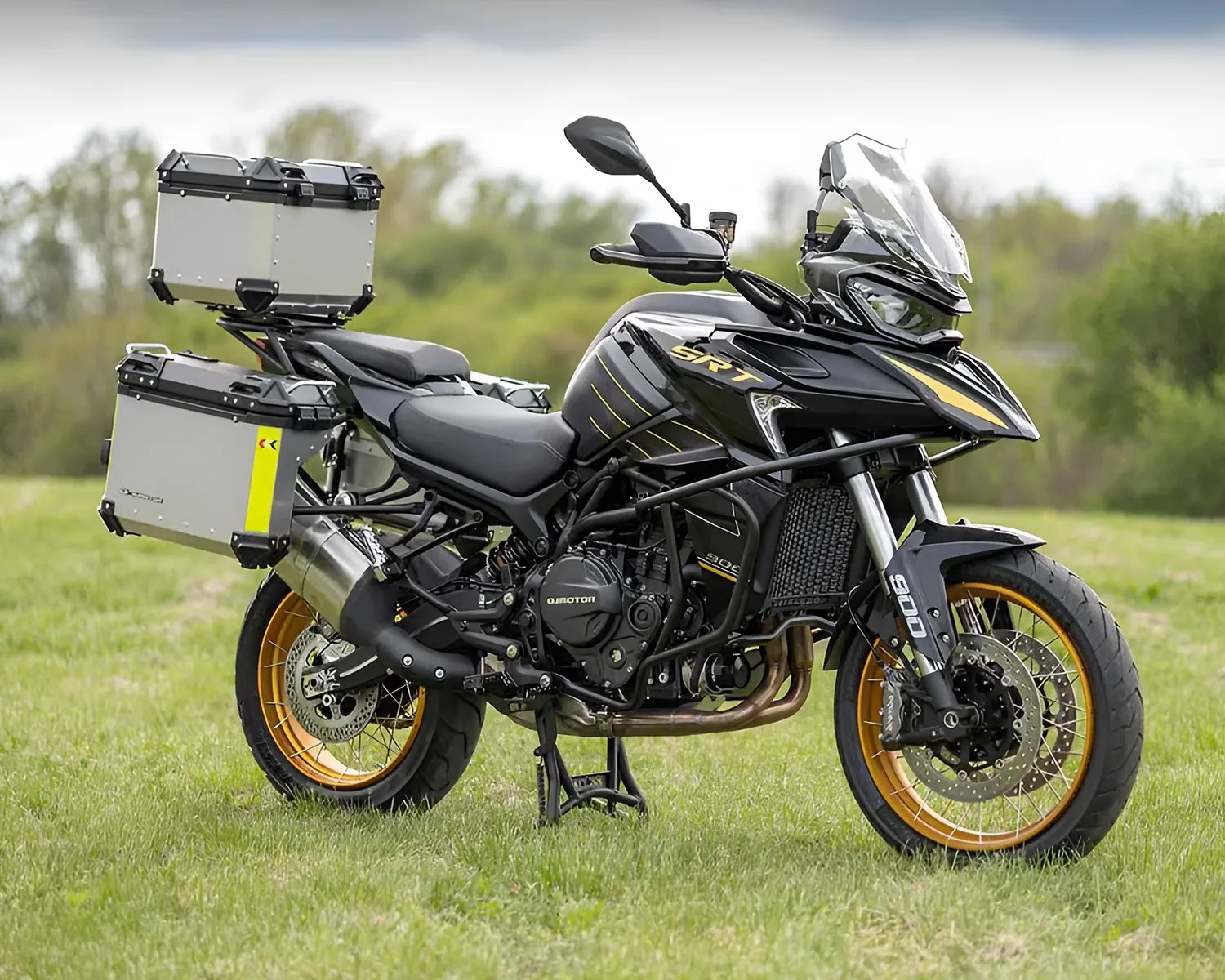 Black and gold adventure motorcycle with three silver storage boxes mounted on the rear and sides, featuring a clear windshield, gold rims, and dual front disc brakes, parked on grass with a blurred green background.
