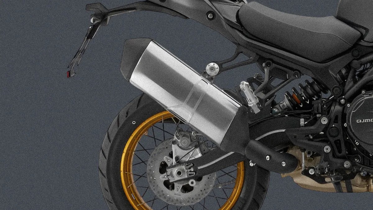 Rear section of a motorcycle featuring a large silver exhaust pipe, a black rear tire with a gold rim, a visible rear suspension spring, and a black seat. The background is plain and dark.