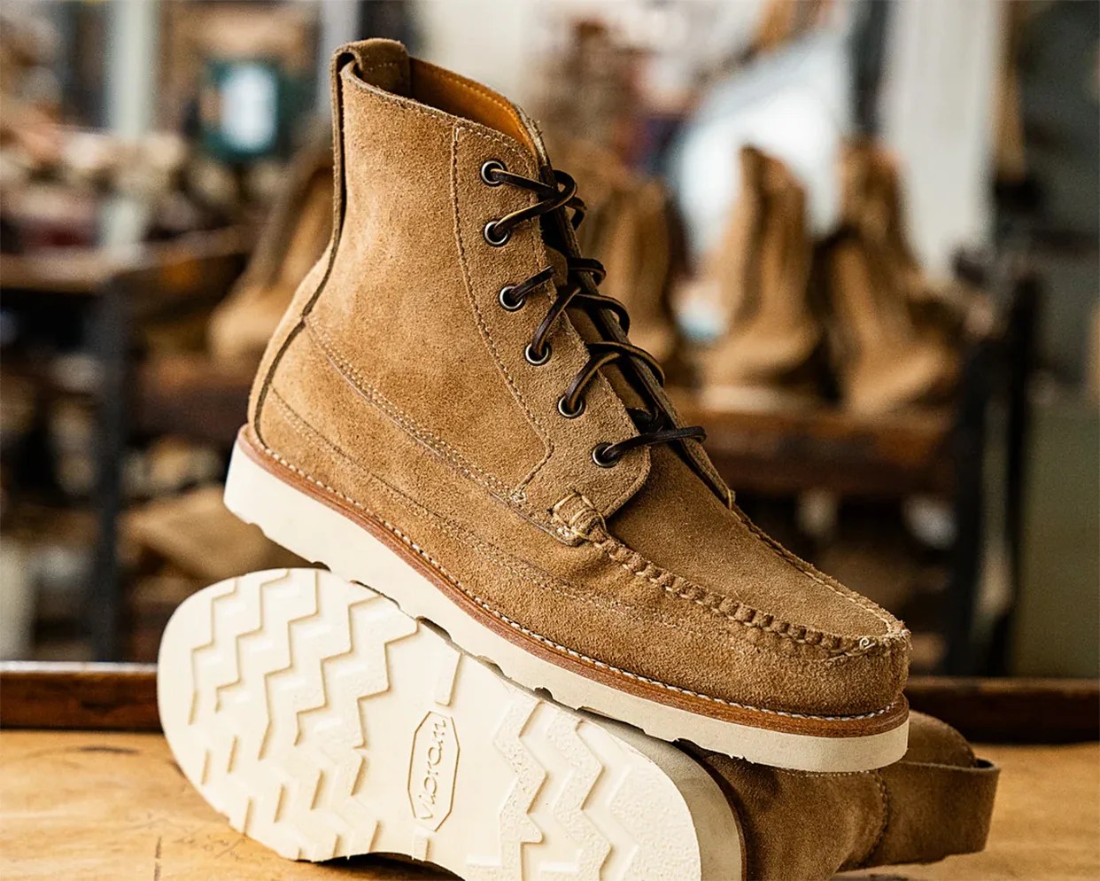 Tan suede moc toe boots with dark brown laces and white Vibram soles, displayed on a wooden surface with a blurred background of similar boots.