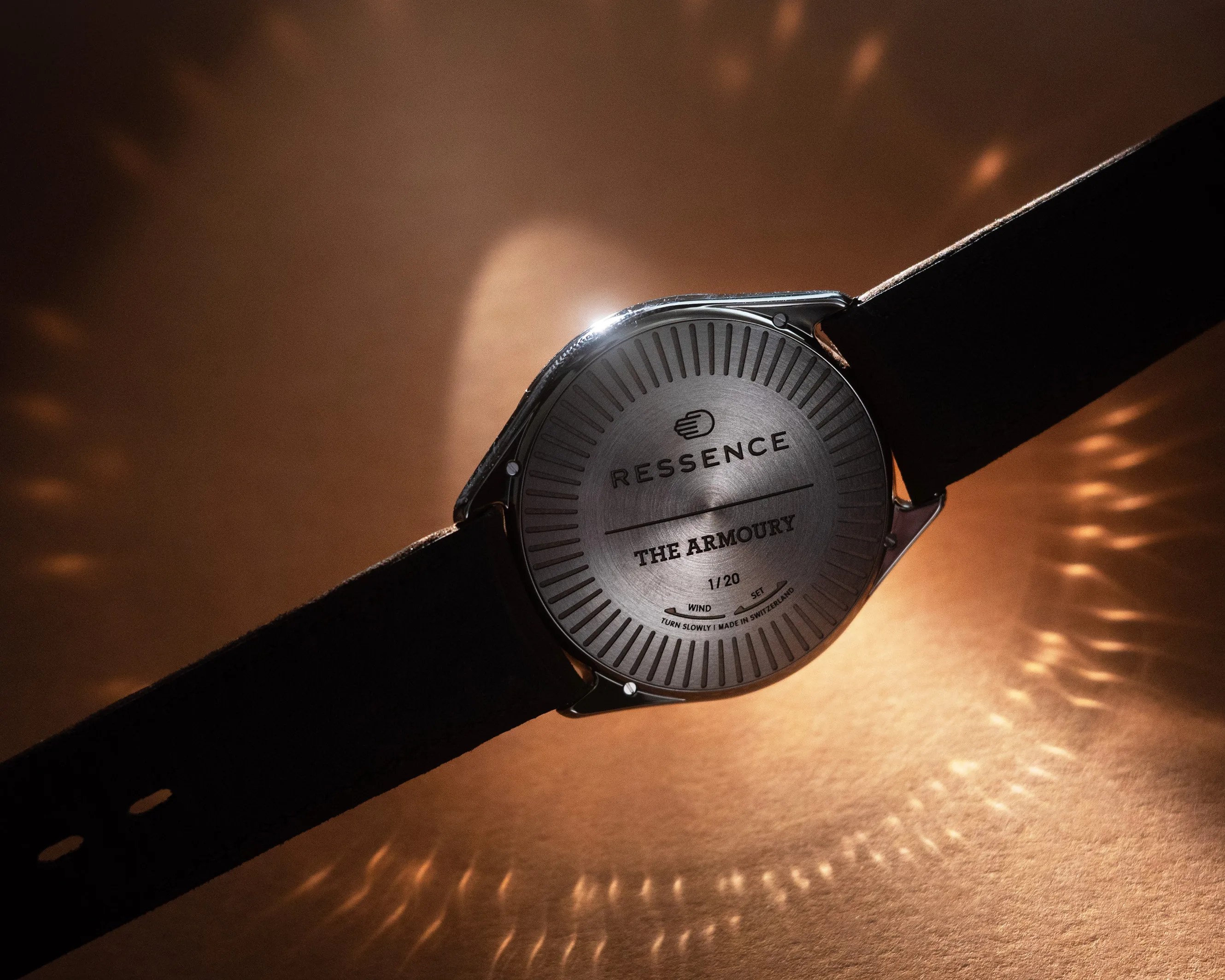 Back view of a Ressence wristwatch with a black leather strap. The watch case is metallic with engraved text reading "RESSENCE," "THE ARMOURY," "1/20," and "TURN SLOWLY | MADE IN SWITZERLAND." The background is warm-toned with light reflections creating a pattern around the watch.