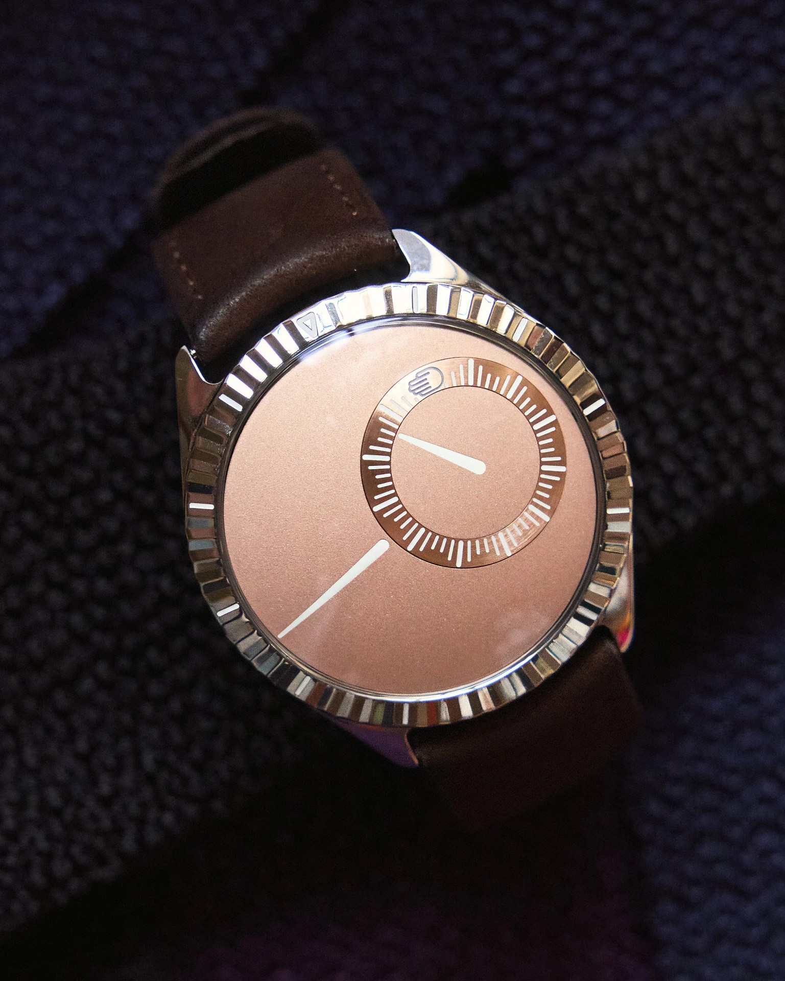 A wristwatch with a rose gold dial and a polished silver bezel featuring ridged edges. The watch has two white hands: a long, slender one on the main dial and a shorter one on a smaller sub-dial positioned near the 2 o'clock area. The watch strap is dark brown leather. The background is a textured dark fabric.