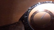 Close-up of a watch face with a textured brown dial, a single hand pointing near the 1 o'clock position, and a black bezel with raised markers. The watch strap appears to be made of brown leather.