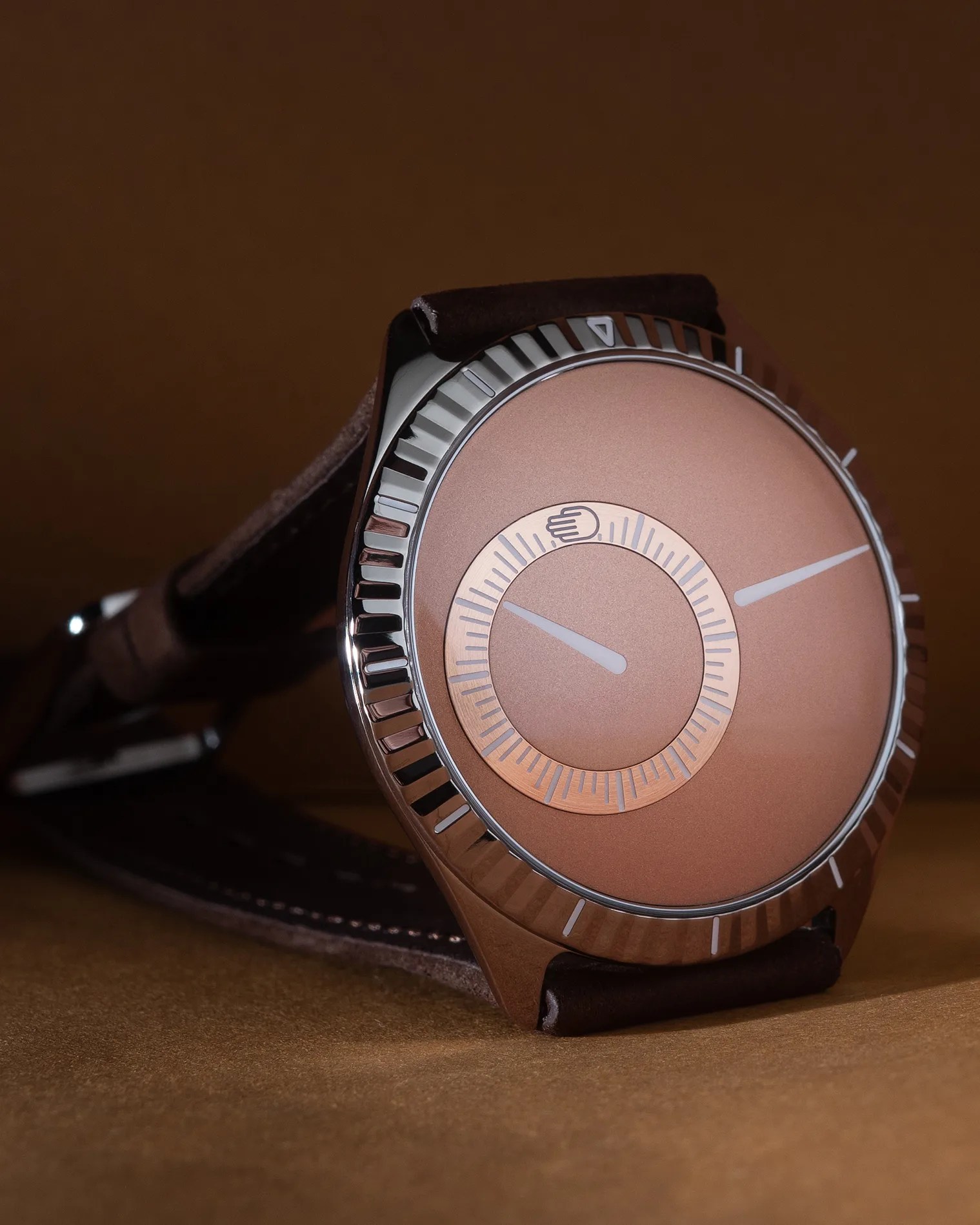 A close-up of a wristwatch with a rose gold dial and a polished silver bezel featuring ridged edges. The watch has a unique design with two white hands, one on the main dial and one on a smaller inner circle marked with minute indicators and a hand icon. The strap appears to be dark brown leather with visible stitching. The background is a matching brown color.