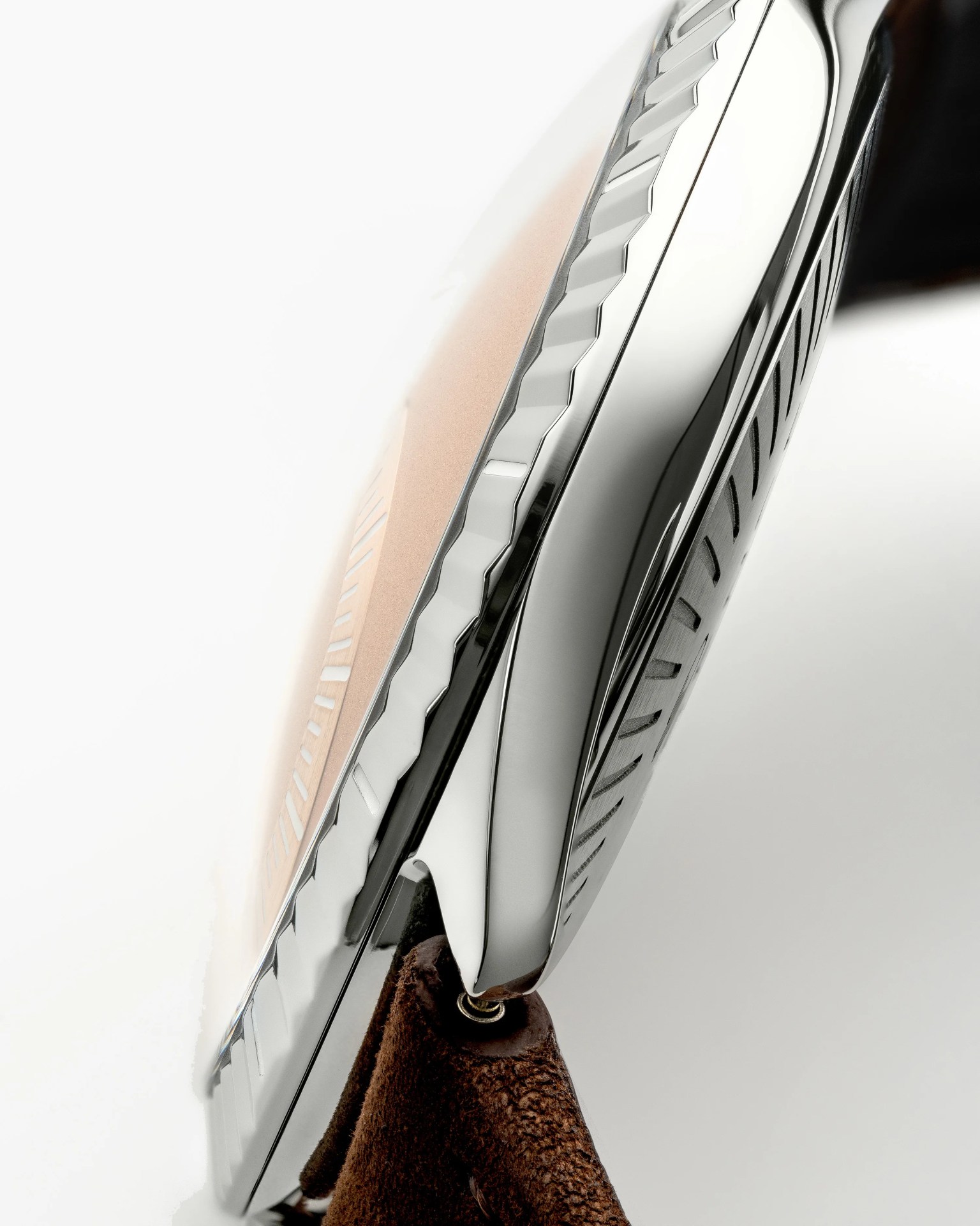 Close-up side view of a polished silver watch case with a ridged bezel and a brown leather strap attached. The watch face is partially visible with a light-colored dial and minimalist hour markers. The background is plain white.