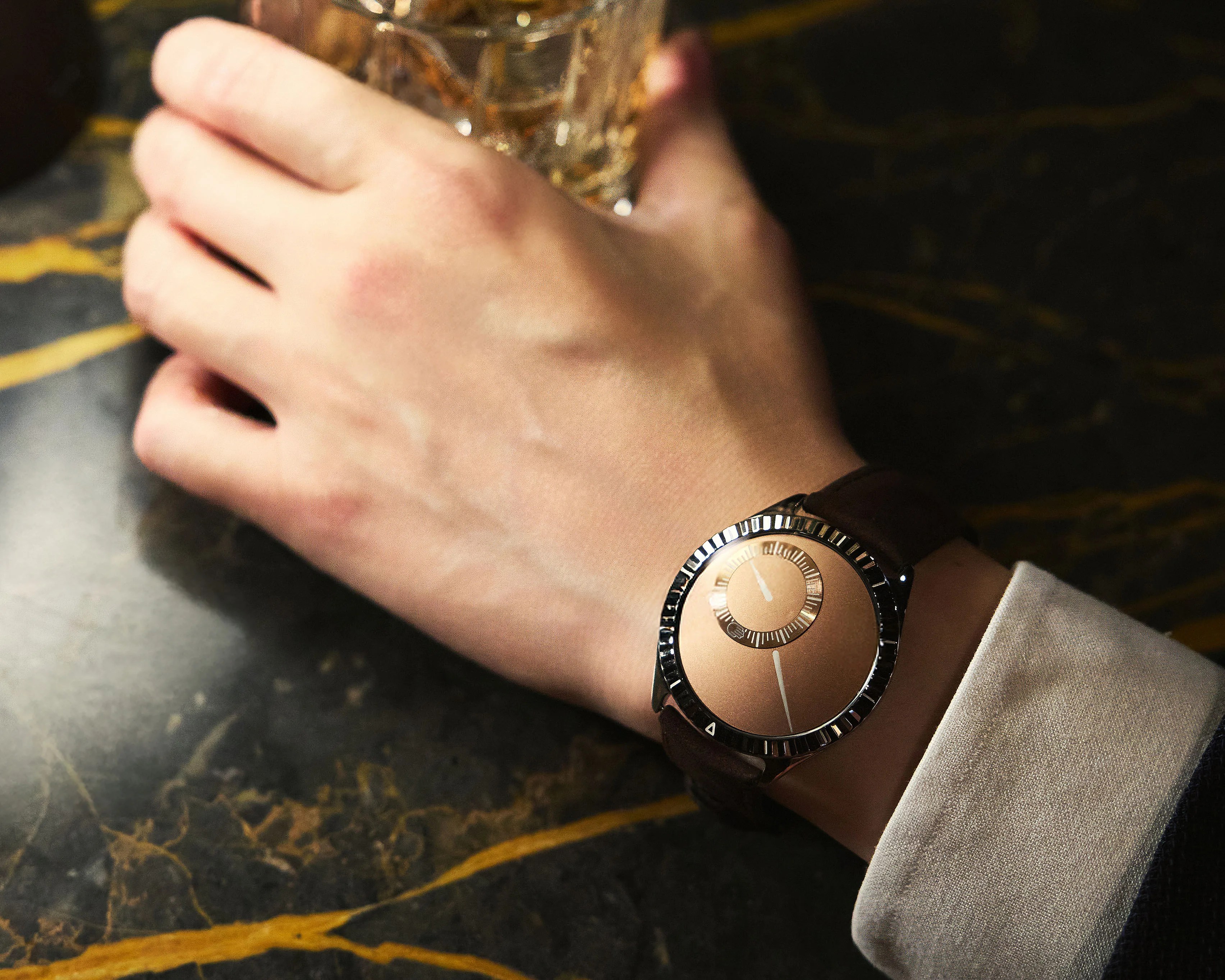 A wristwatch with a minimalist design featuring a round, metallic rose gold dial and a smaller inner circle with hour markers. The watch has a dark brown leather strap and a shiny, ridged bezel. The hand wearing the watch is holding a glass with a textured pattern, resting on a dark marble surface with yellowish veins. The person is wearing a white shirt cuff.