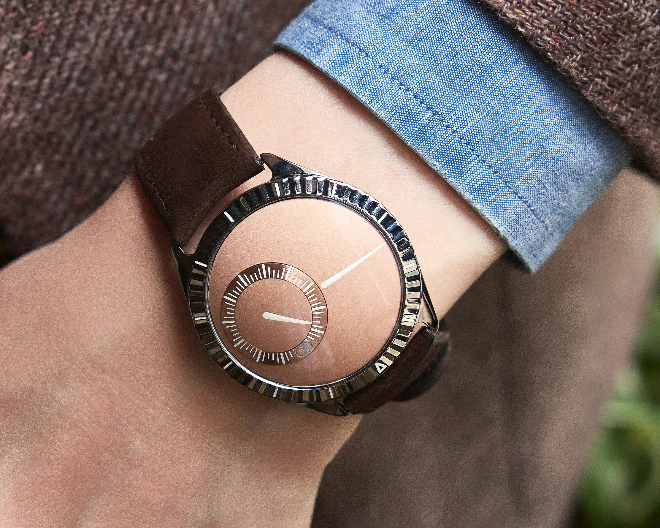 Wristwatch with a minimalist design featuring a rose gold dial, no numbers, and two white hands—one on the main dial and one on a smaller sub-dial. The watch has a dark brown leather strap and a polished silver bezel with ridged edges. The wearer is dressed in a blue shirt and a brown textured sweater.