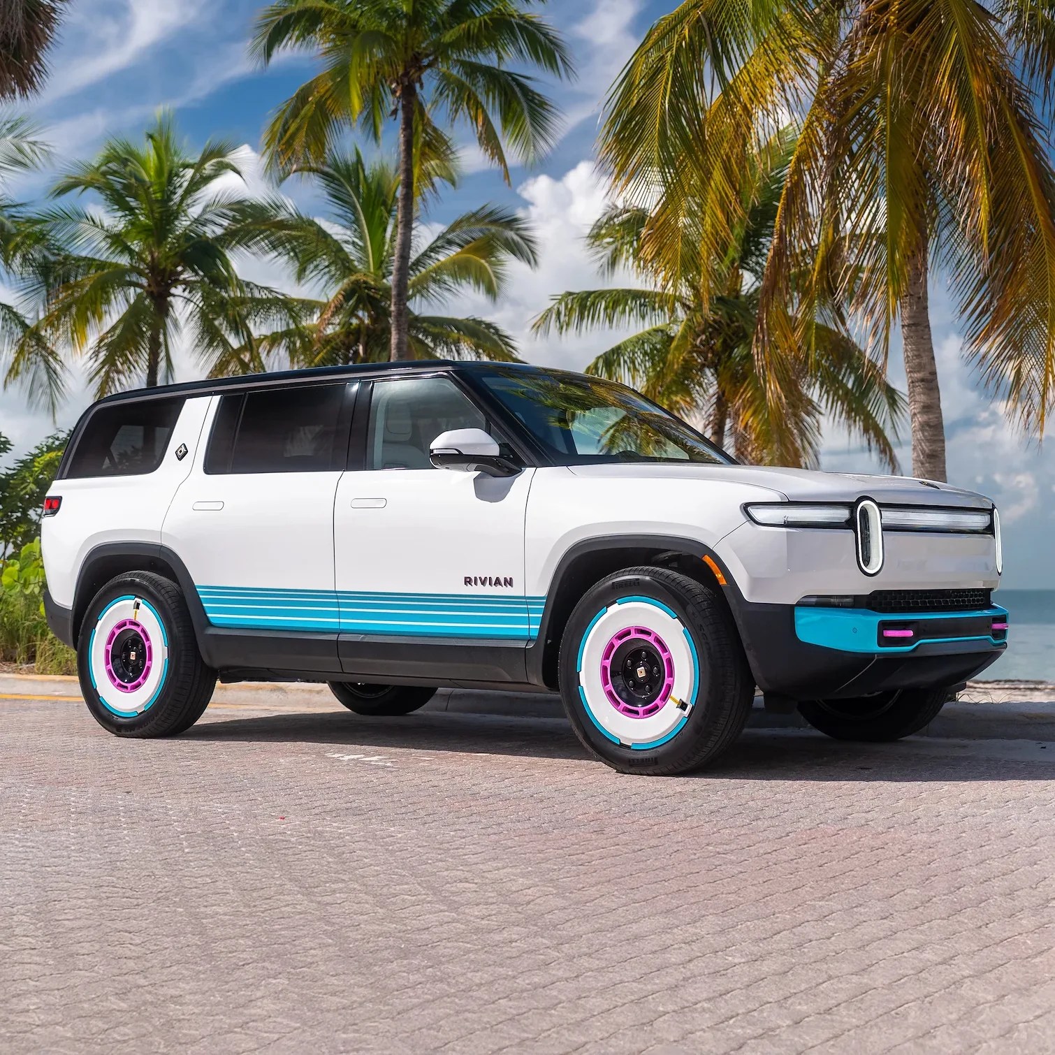 White Rivian SUV with black roof and blue and pink accents on the wheels and lower side panels, parked on a paved surface with palm trees and a partly cloudy sky in the background.