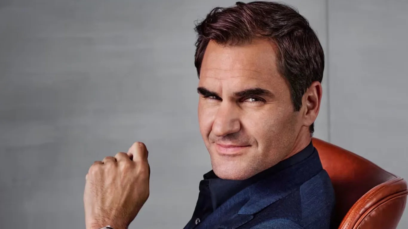 Roger Federer is seated in a brown leather chair, wearing a dark blue suit jacket and a black shirt, looking directly at the camera.
