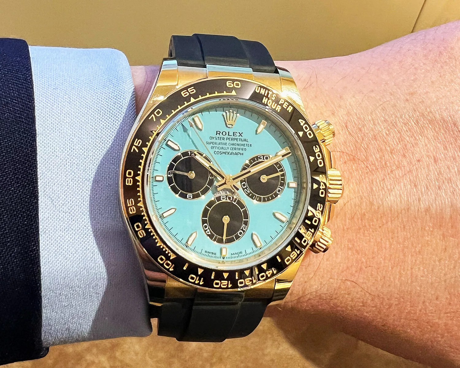 Rolex Oyster Perpetual Cosmograph Daytona wristwatch with a gold case, black bezel marked with units per hour, and a light blue dial featuring three black subdials. The watch has gold hour markers and hands, and is paired with a black rubber strap. The watch is worn on a wrist with a light blue shirt cuff and dark jacket sleeve visible.