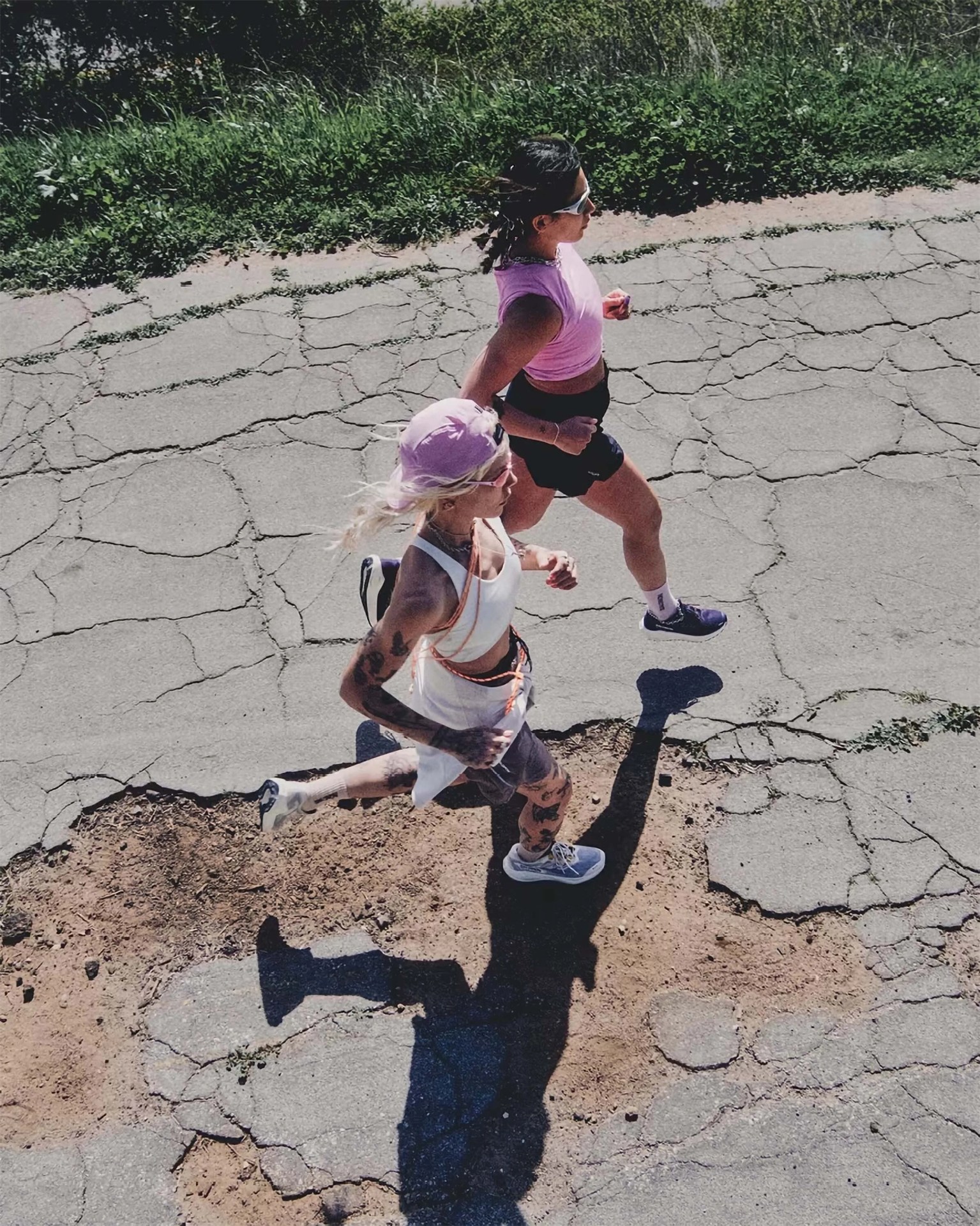Two women are running side by side on a cracked and uneven paved surface with patches of dirt. The woman on the left has light hair, wears a light purple cap, white sportswear, and sneakers, and has visible tattoos on her arms and legs. The woman on the right has dark hair tied back, wears sunglasses, a pink sleeveless top, black shorts, white socks, and dark running shoes. There is green grass bordering the pavement at the top of the image.