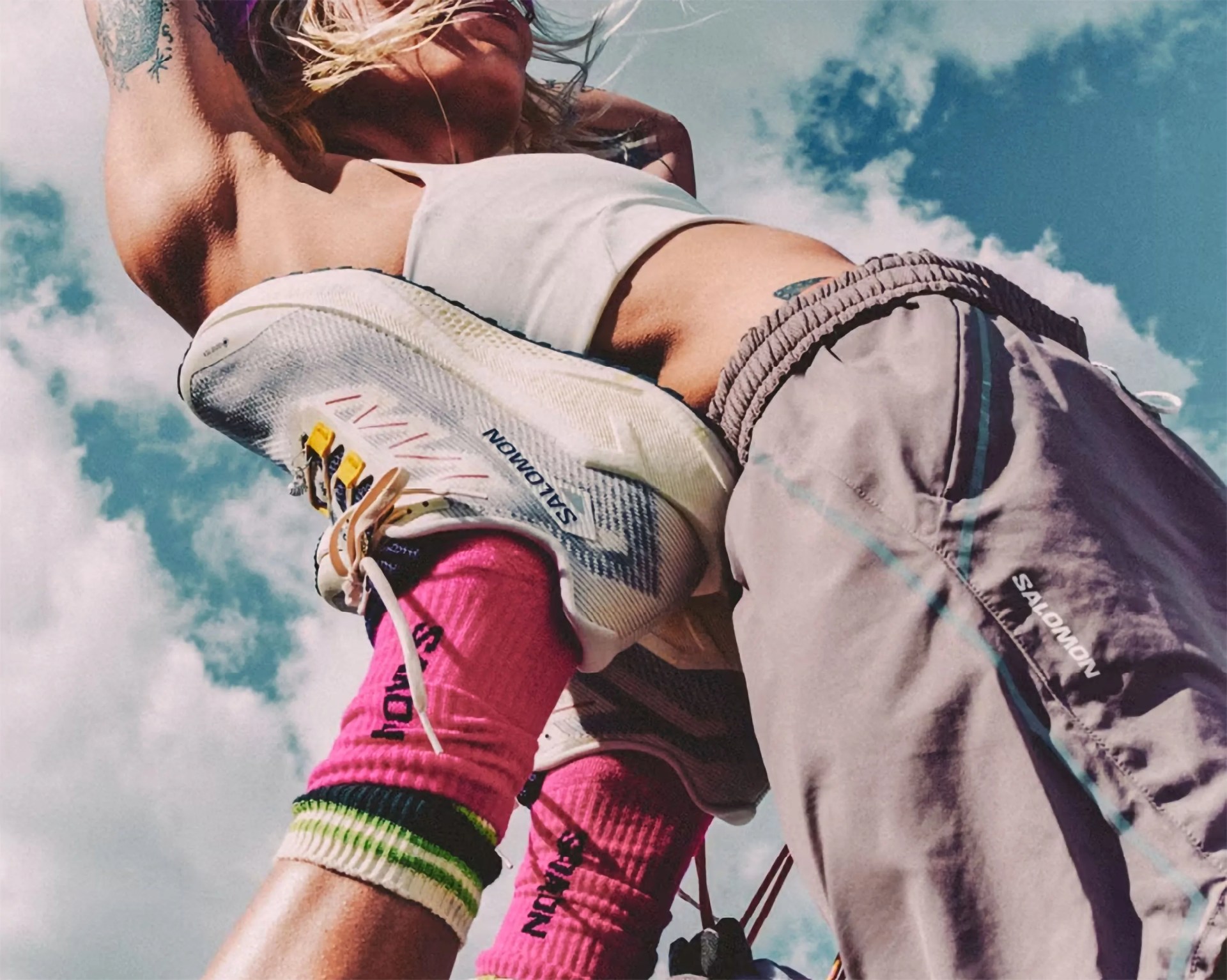 A person wearing light gray Salomon pants and white Salomon sneakers with pink socks featuring black text. The photo is taken from a low angle against a partly cloudy blue sky, emphasizing the footwear and lower body. The person has a tattoo on their upper arm and is wearing a white crop top.