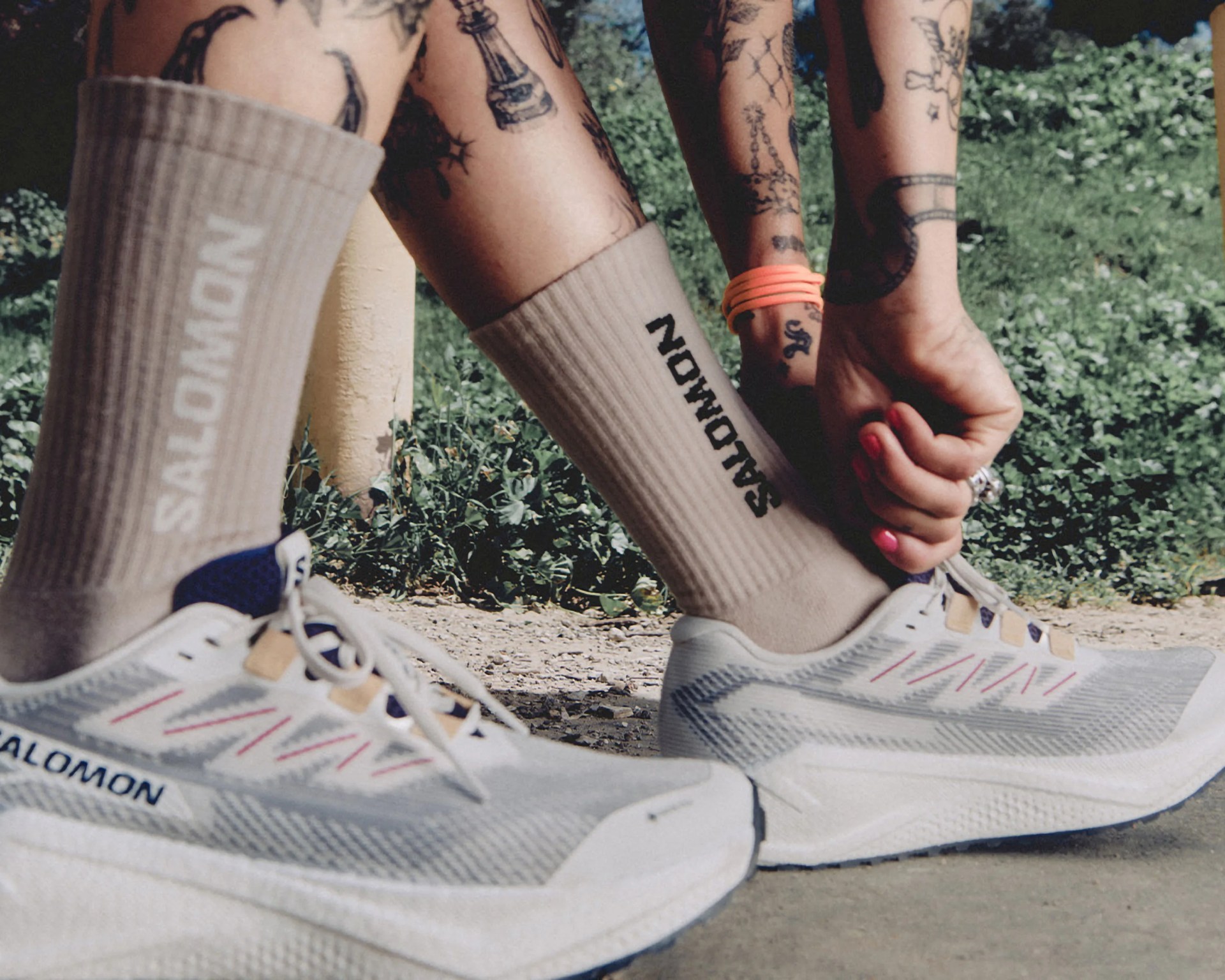 Close-up of a person wearing beige Salomon socks and white Salomon running shoes with blue and tan accents, tying the laces on one shoe. The person's legs are tattooed, and they have pink nail polish on their fingernails and an orange hair tie on their wrist. The background shows green foliage and a dirt path.
